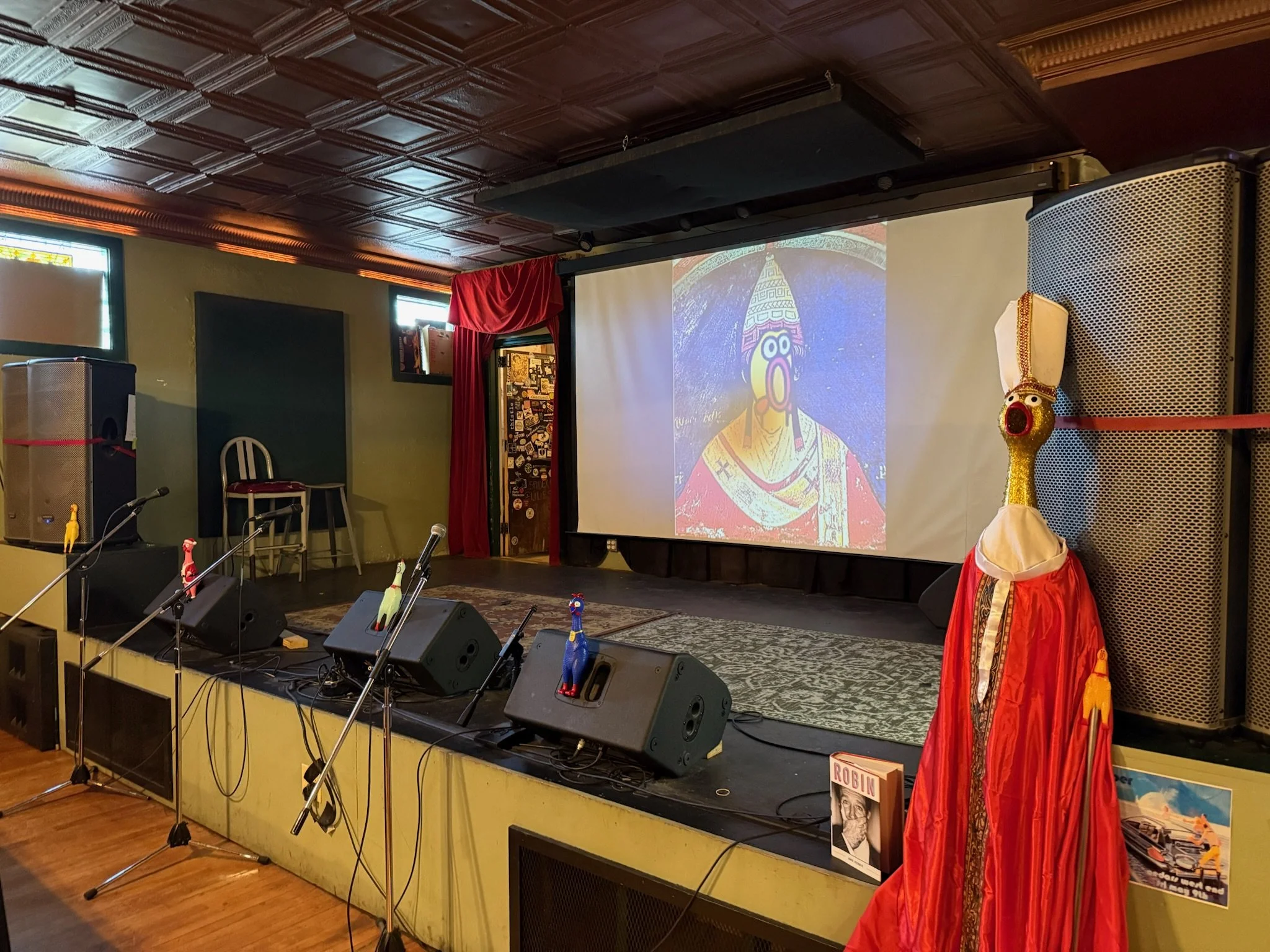 Stage with a large projection screen displaying a colorful drawing of a person with a bird-like beak, wearing traditional attire. The stage has a large puppet resembling a bird with a long neck, white head, and wearing a red dress. There are microphones with small bird-like figures on the stands in front of the stage. The room has a wooden ceiling, small windows, and a green wall.