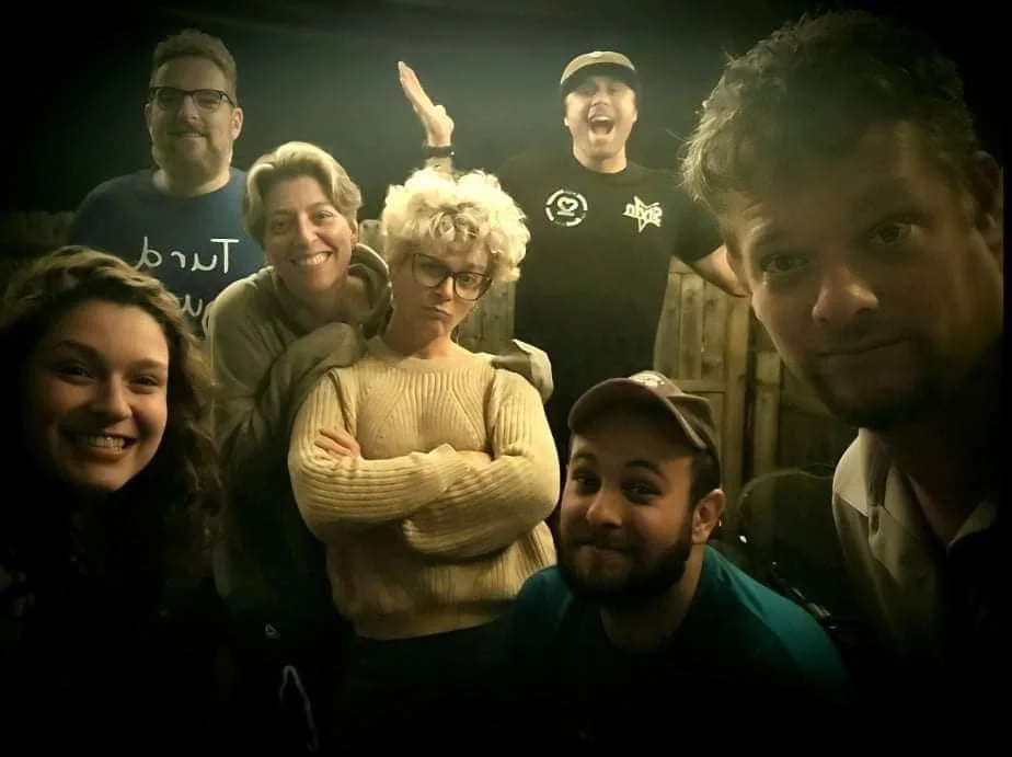 Group of seven people posing together indoors, smiling and making playful expressions, with a dark background and some wooden panels.