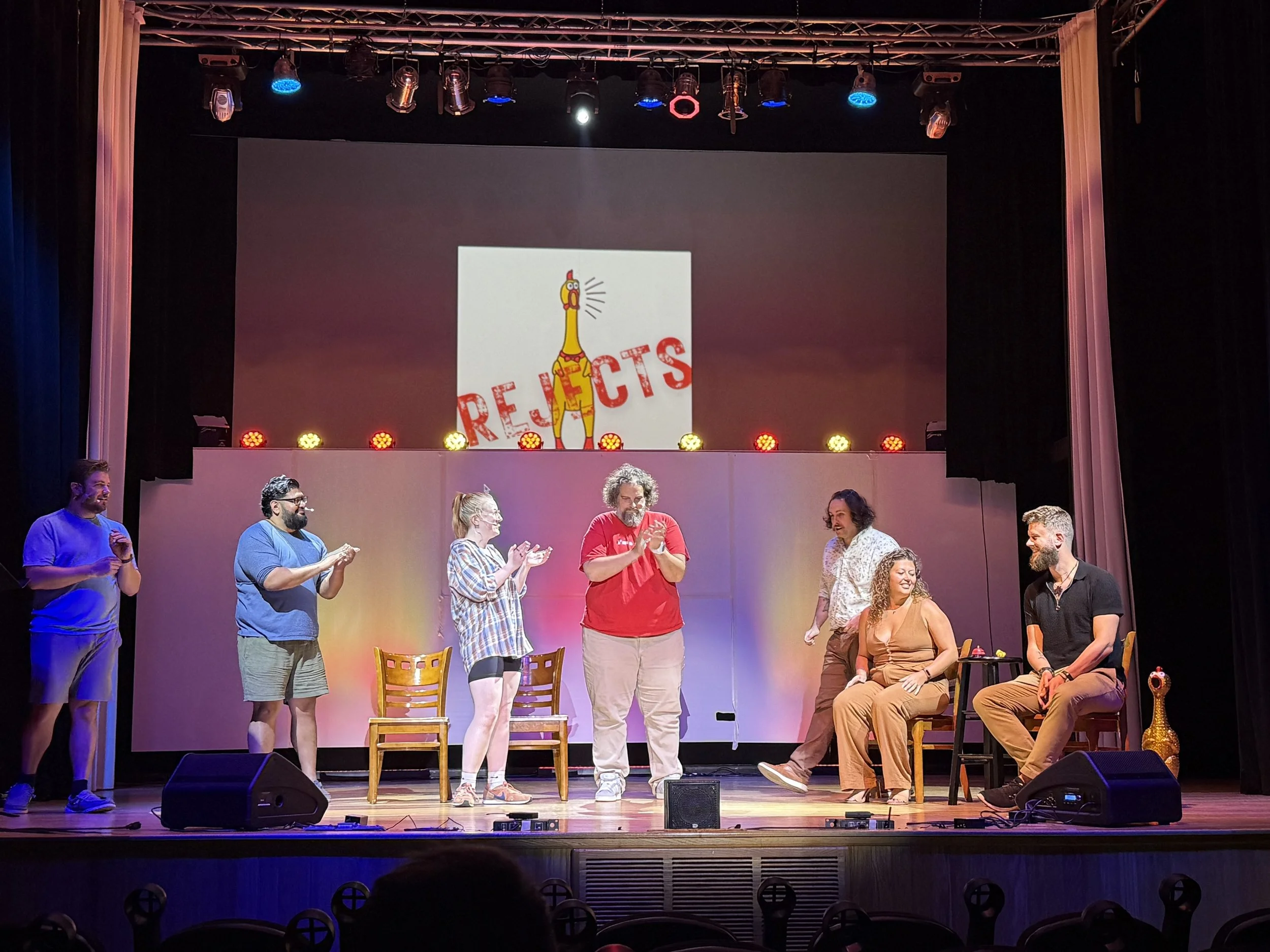 A group of seven people on a stage during a theatrical performance or improvisation event, with a backdrop featuring a cartoon giraffe and the word 'REJECTS' in bold red letters. Some characters are standing, others are seated, engaging in conversation or laughter, with colorful stage lights overhead.
