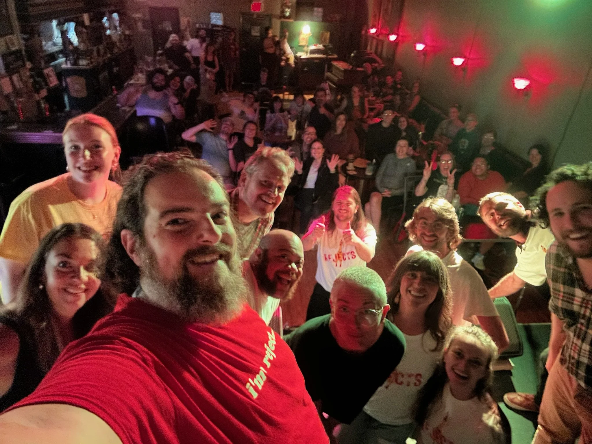 Group selfie of comedians and audience members at a comedy club, smiling and posing together on stage, with a packed audience in the background.