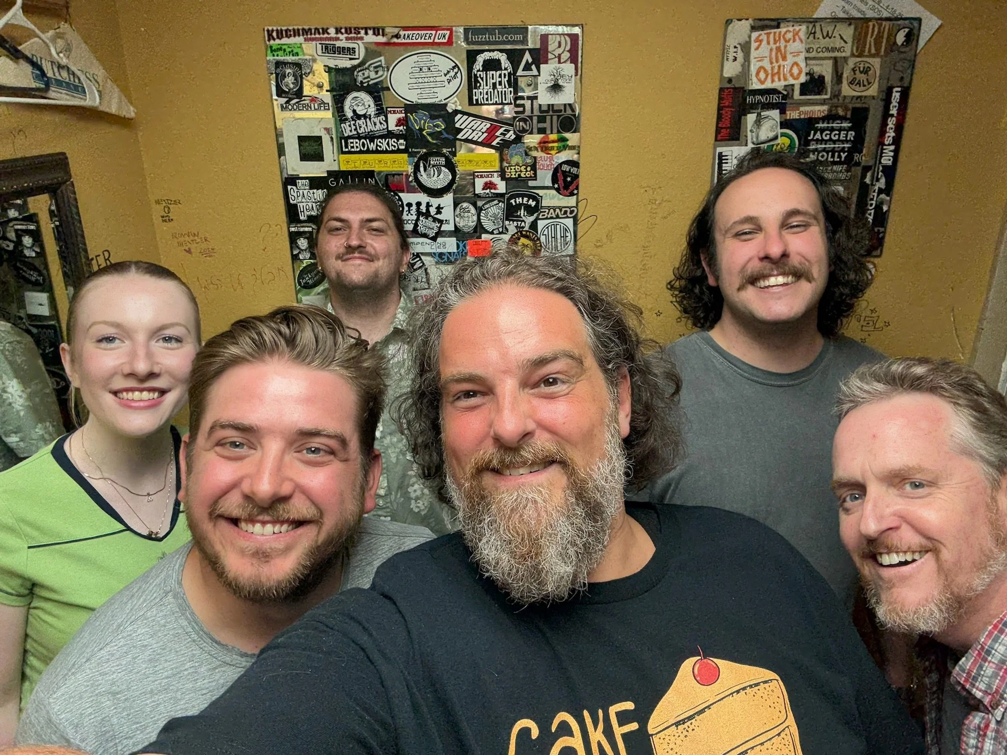 Group of six people smiling for a selfie in front of a wall decorated with patches and stickers.
