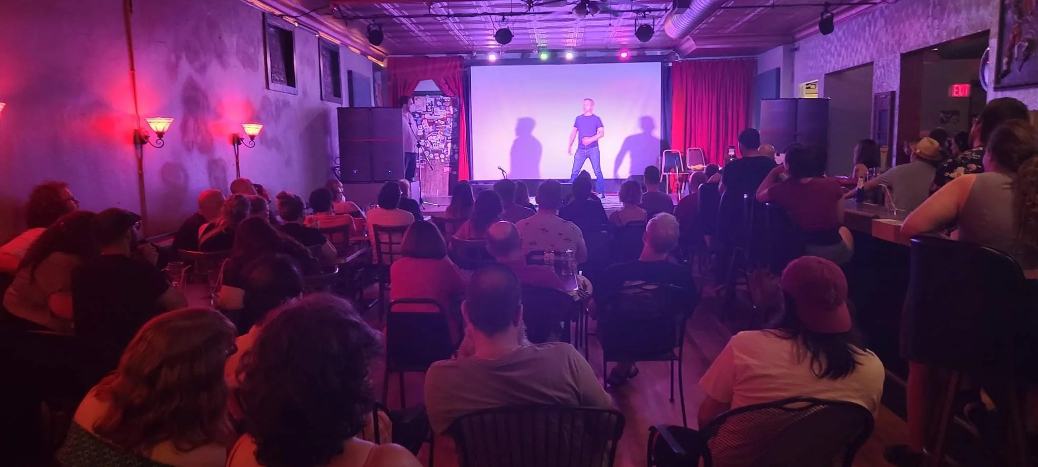 Audience watching a stand-up comedy show on a stage in a dimly lit room with purple and red lighting.