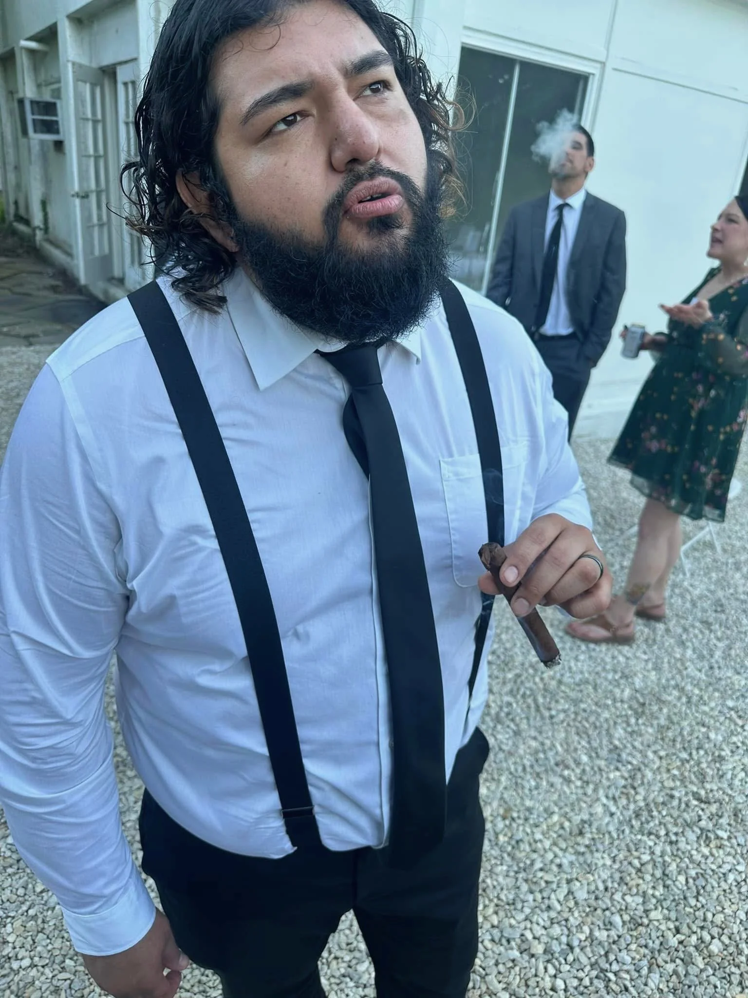 Man with dark, curly hair and beard in white shirt, black tie, and suspenders holding a cigar, standing outdoors with gravel ground, with background of three people, two in suits and a woman in a floral dress, one of whom is smoking and the woman is holding a cup.