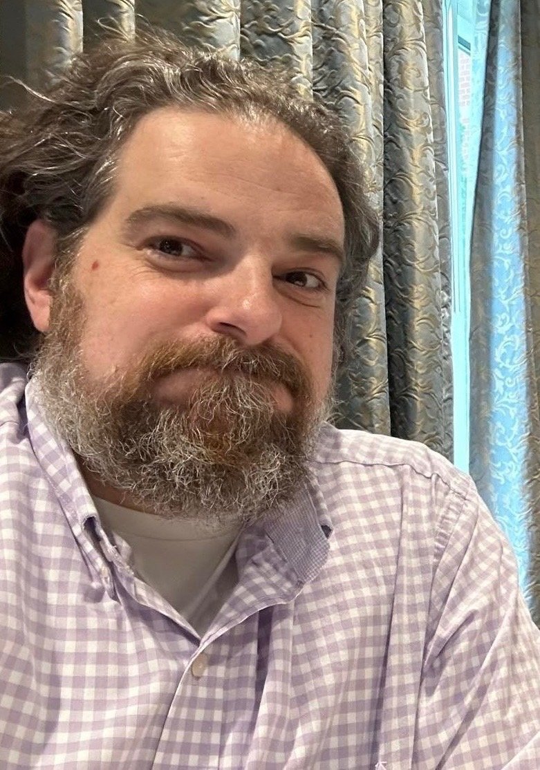 A man with a beard and sideswept hair, wearing a purple checkered shirt, sitting in front of curtains with a window on the right side.
