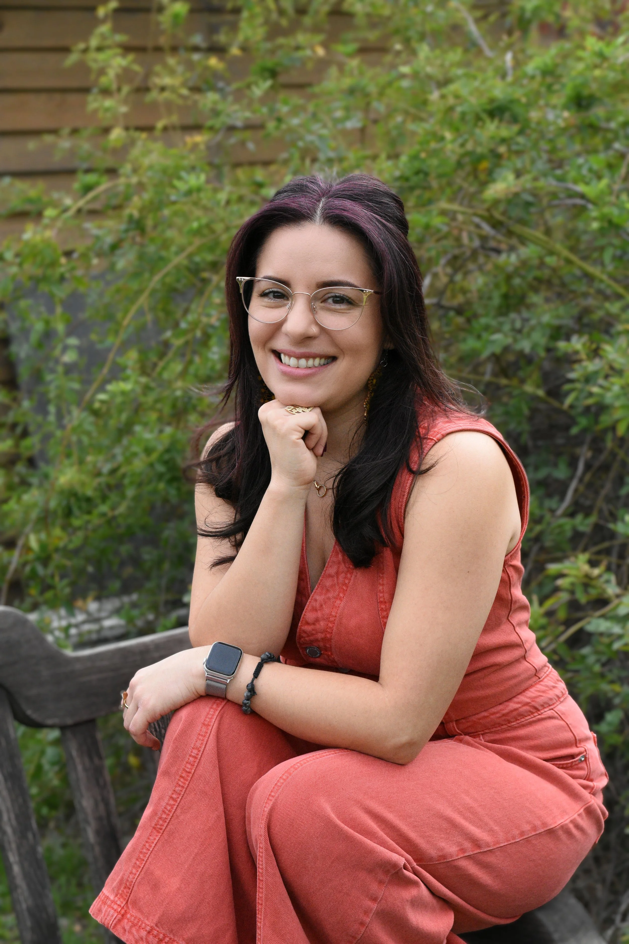 Therapist Lili smiling with hand holding her chin on a chair in relaxed position with greenery outdoor setting