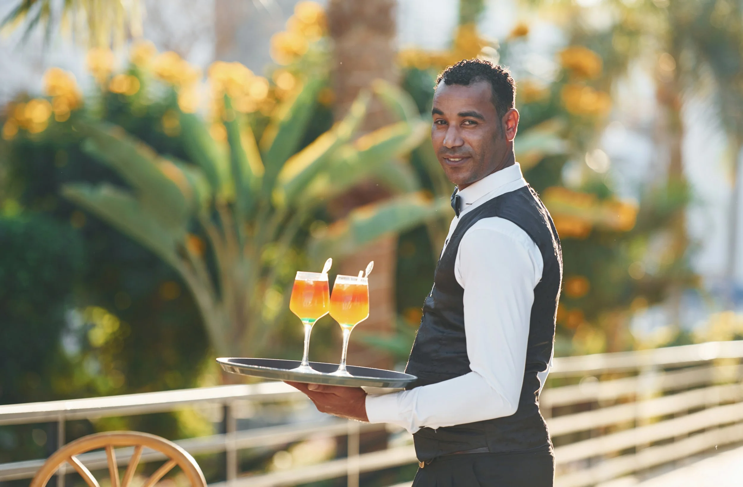 A smiling waiter in a black vest and white shirt holds a tray with two tropical drinks in tall glasses, outdoors with green plants and trees in the background.