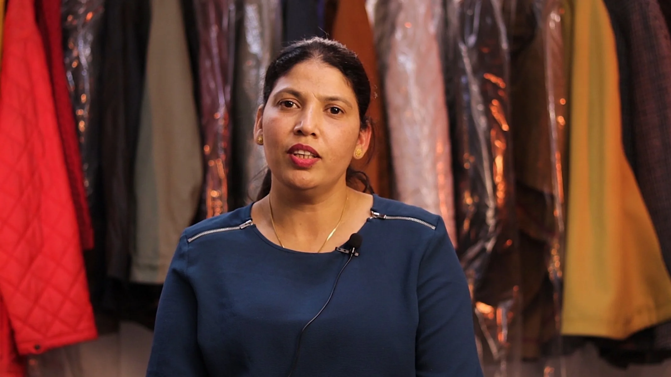 A woman with dark hair is speaking, wearing a blue shirt and a lapel microphone, with colorful hanging garments in the background.