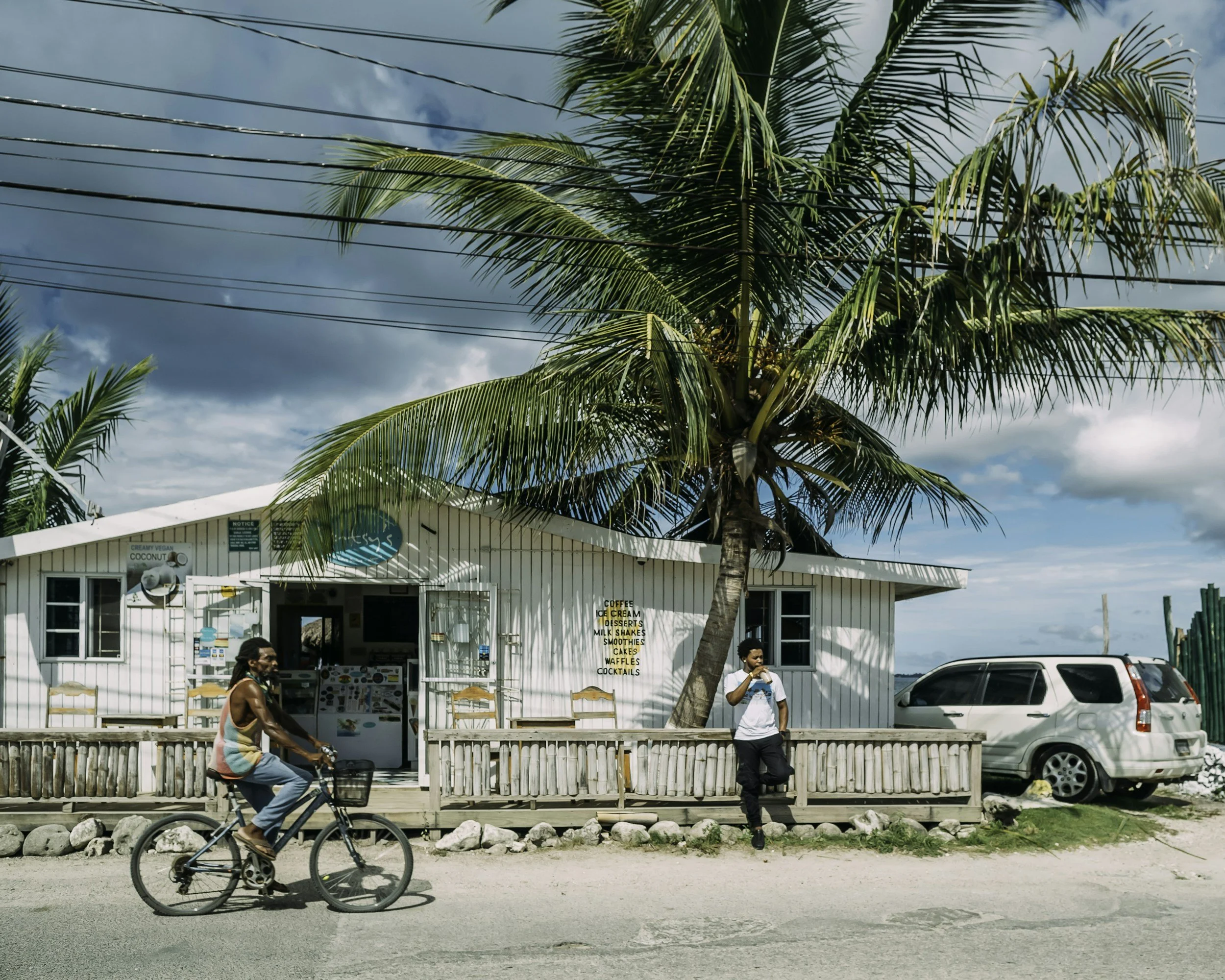 A small white beachside cafe with a sign listing coffee, ice cream, desserts, smoothies, cakes, waffles, and cocktails, situated under a large palm tree with two people outside: one riding a bicycle and the other leaning against a wooden railing, with a car parked nearby and a partly cloudy sky overhead.