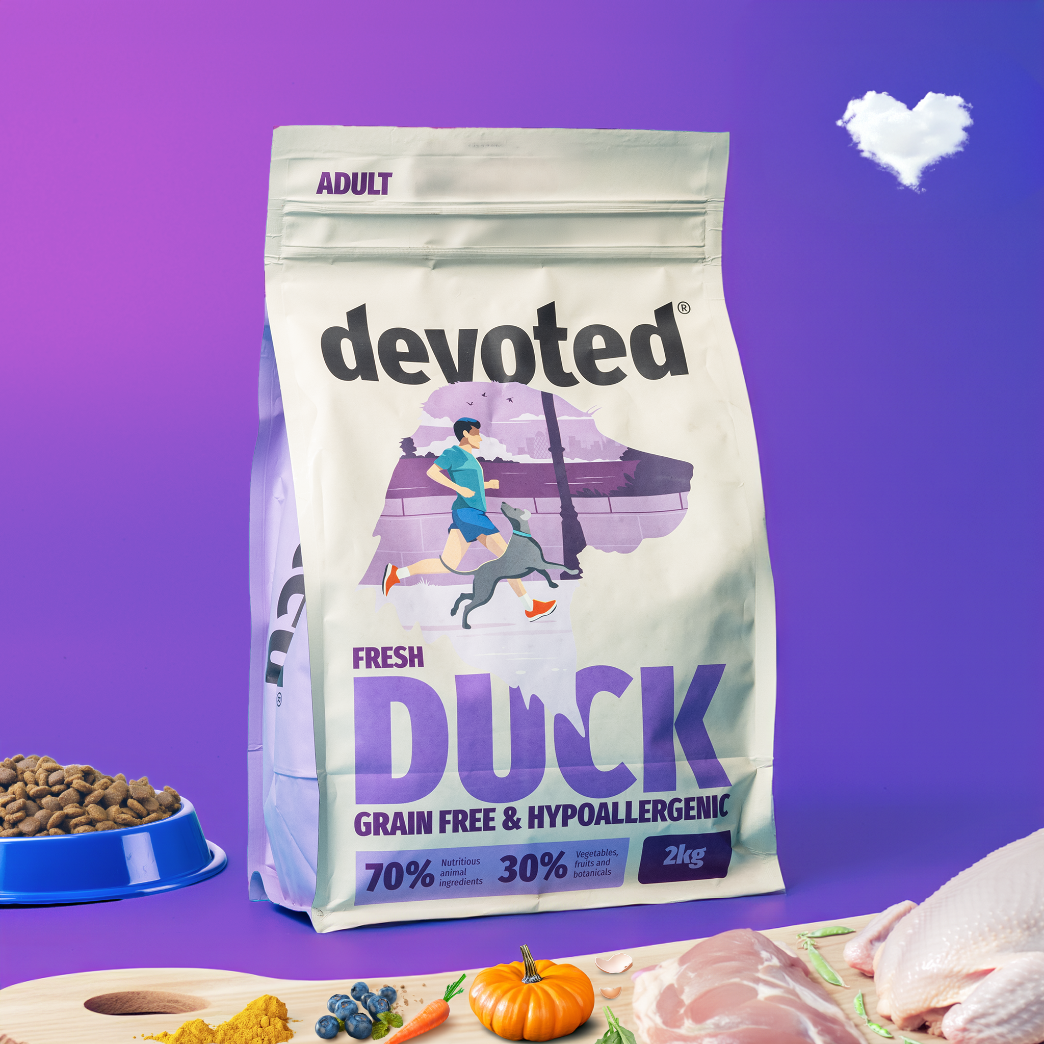 A 2kg bag of devoted brand dog food labeled as adult, fresh duck, grain-free, hypoallergenic, and with 70% animal ingredients and 30% vegetables, fruits, and botanicals. The bag features an illustration of a person running with a dog in front of a purple and white background. Next to the bag, there is a blue bowl of dry dog food, some raw meat, a pumpkin, blueberries, a carrot, and some greens placed on a wooden surface.