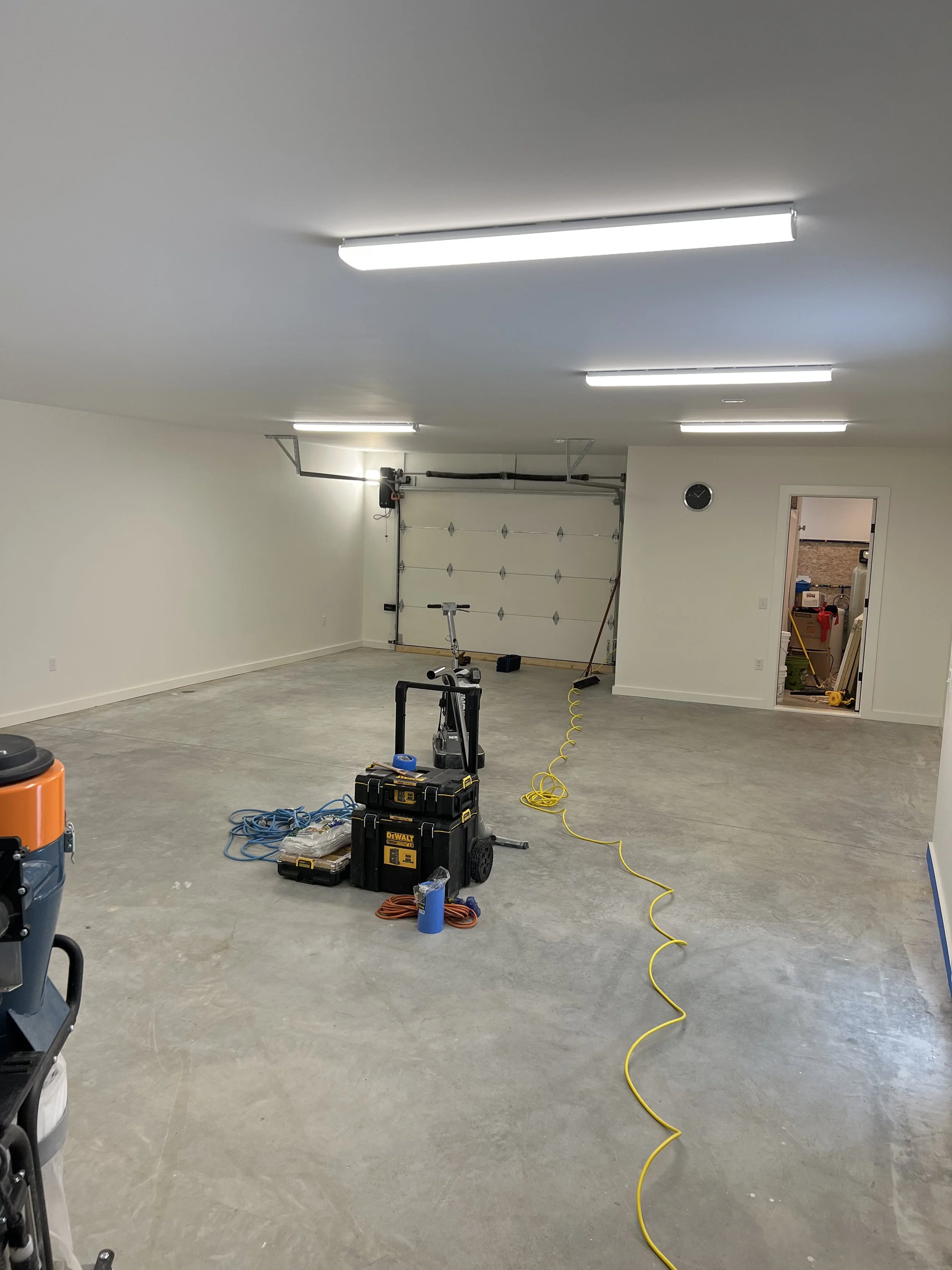 Empty garage with fluorescent lighting, open garage door, tools and equipment on the concrete floor, yellow extension cord, and a doorway leading into another room.