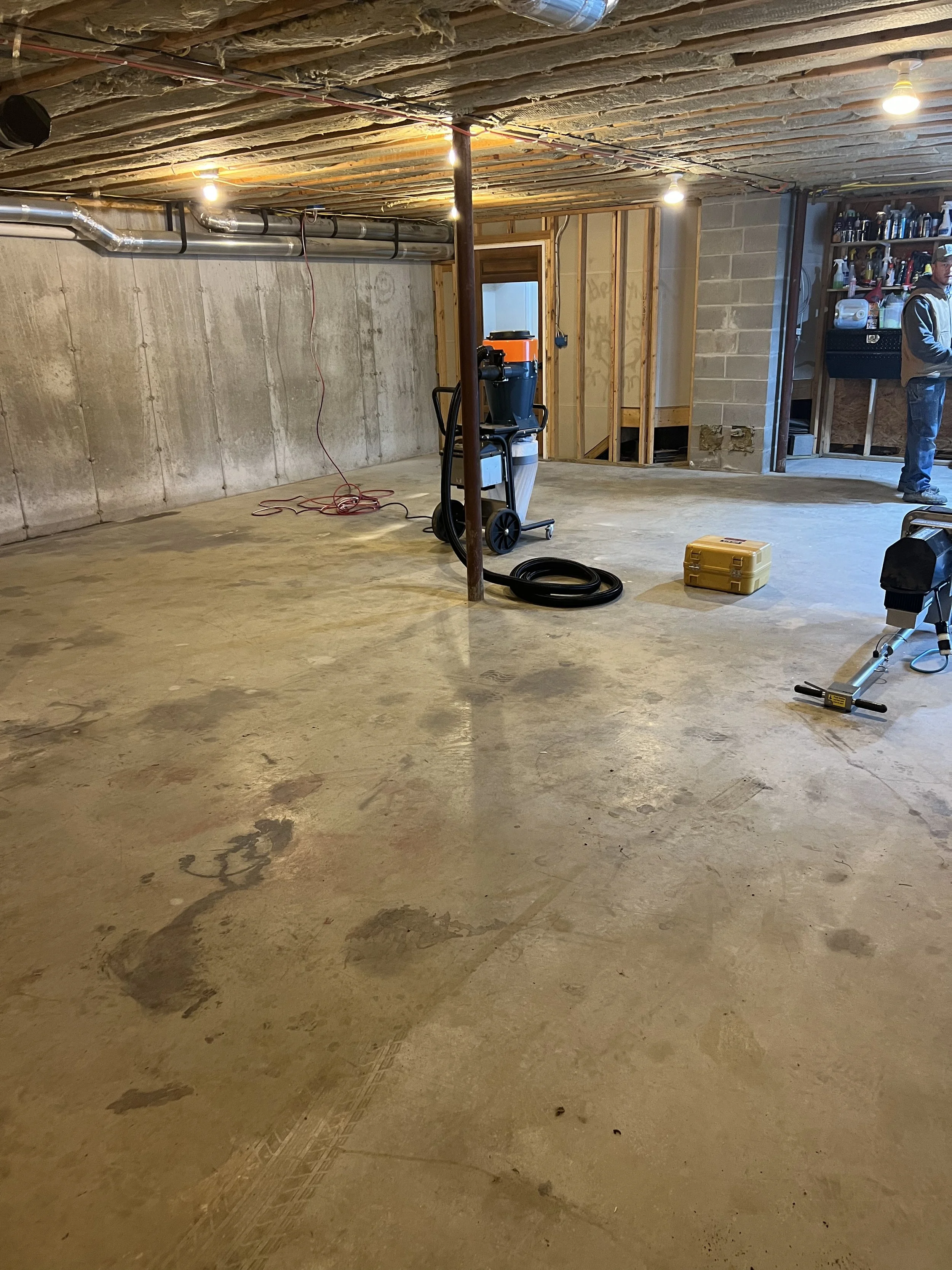 An unfinished basement with exposed ceiling beams, ductwork, and concrete floors. Tools and equipment are scattered around, including a wet/dry vacuum, toolbox, and construction materials. A person stands near a workbench in the background.