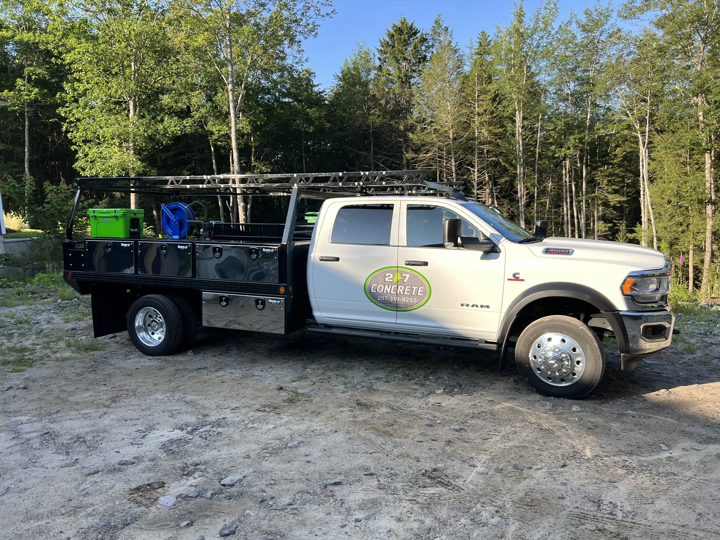 A white utility truck with a black flatbed, parked outdoors on a dirt area with trees in the background. The truck has logo for 24/7 Concrete on the door and construction equipment on the flatbed.