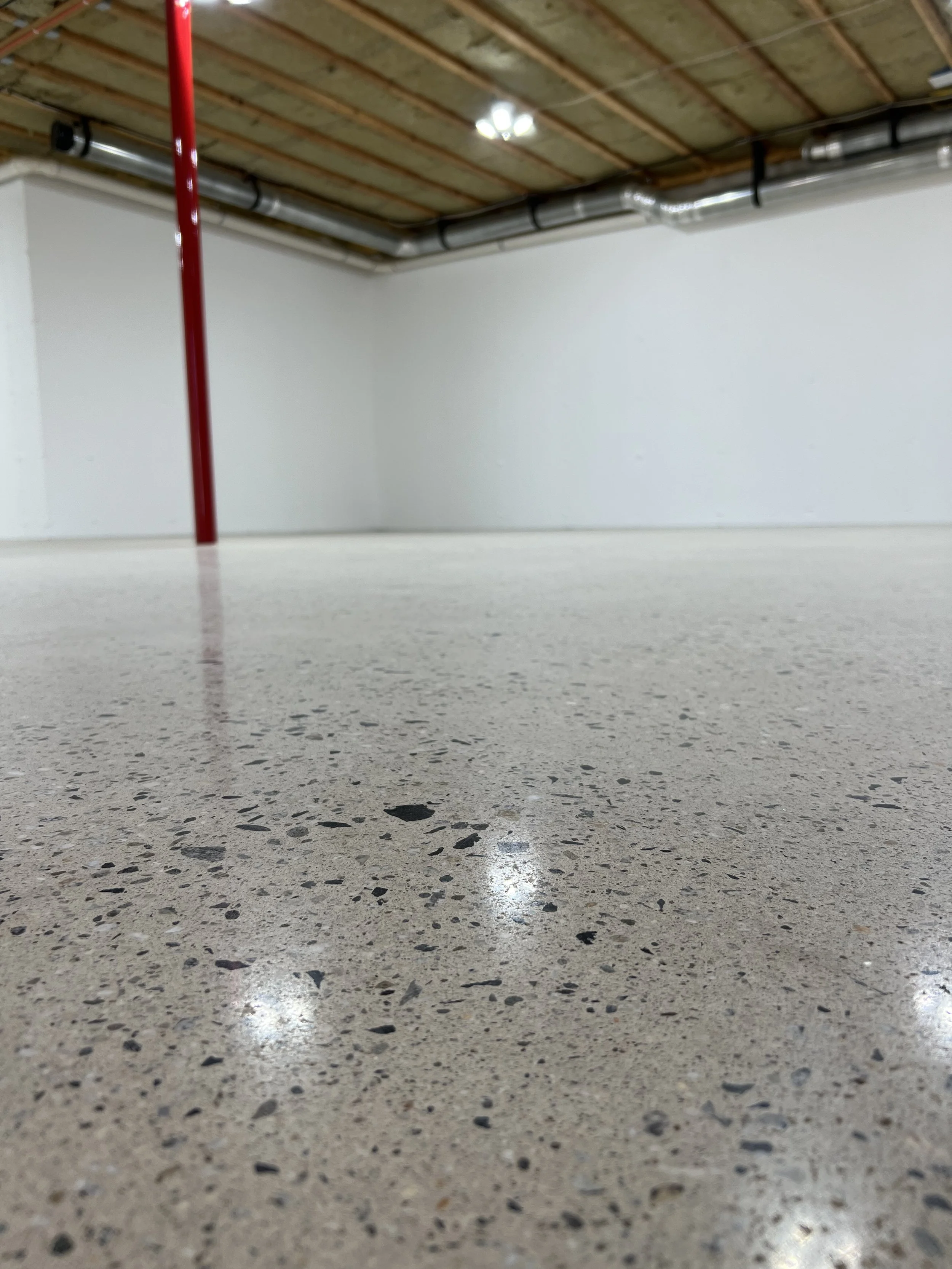 Empty basement or parking garage with polished concrete floor, white painted walls, and visible ductwork on the ceiling. A red support pole is present in the foreground.