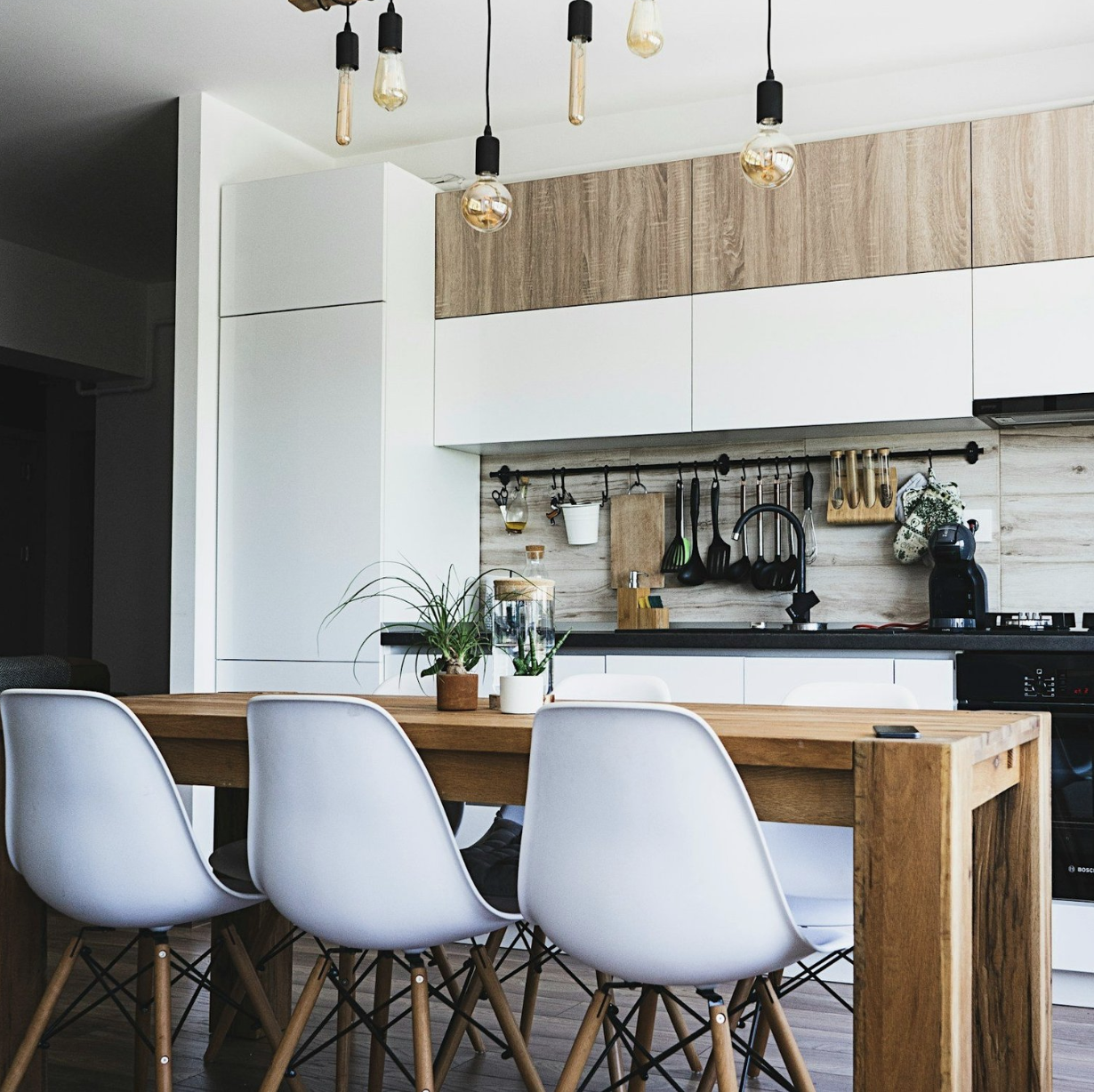 Modern kitchen with wooden table, white chairs, pendant light fixtures, wall-mounted utensils, and a plant on the table.