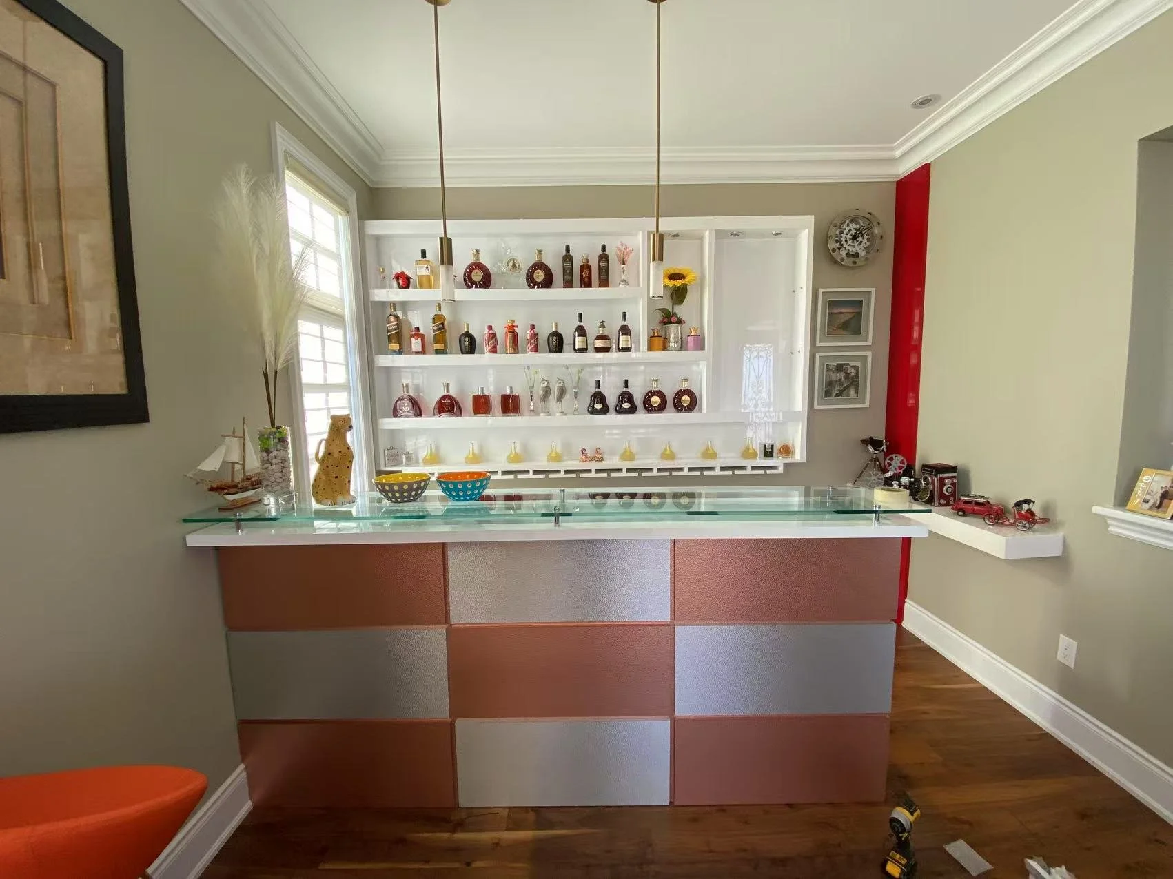 A home bar with a glass countertop, colorful bowls, decorative bottles on white shelves, and small collectible cars on a white ledge.