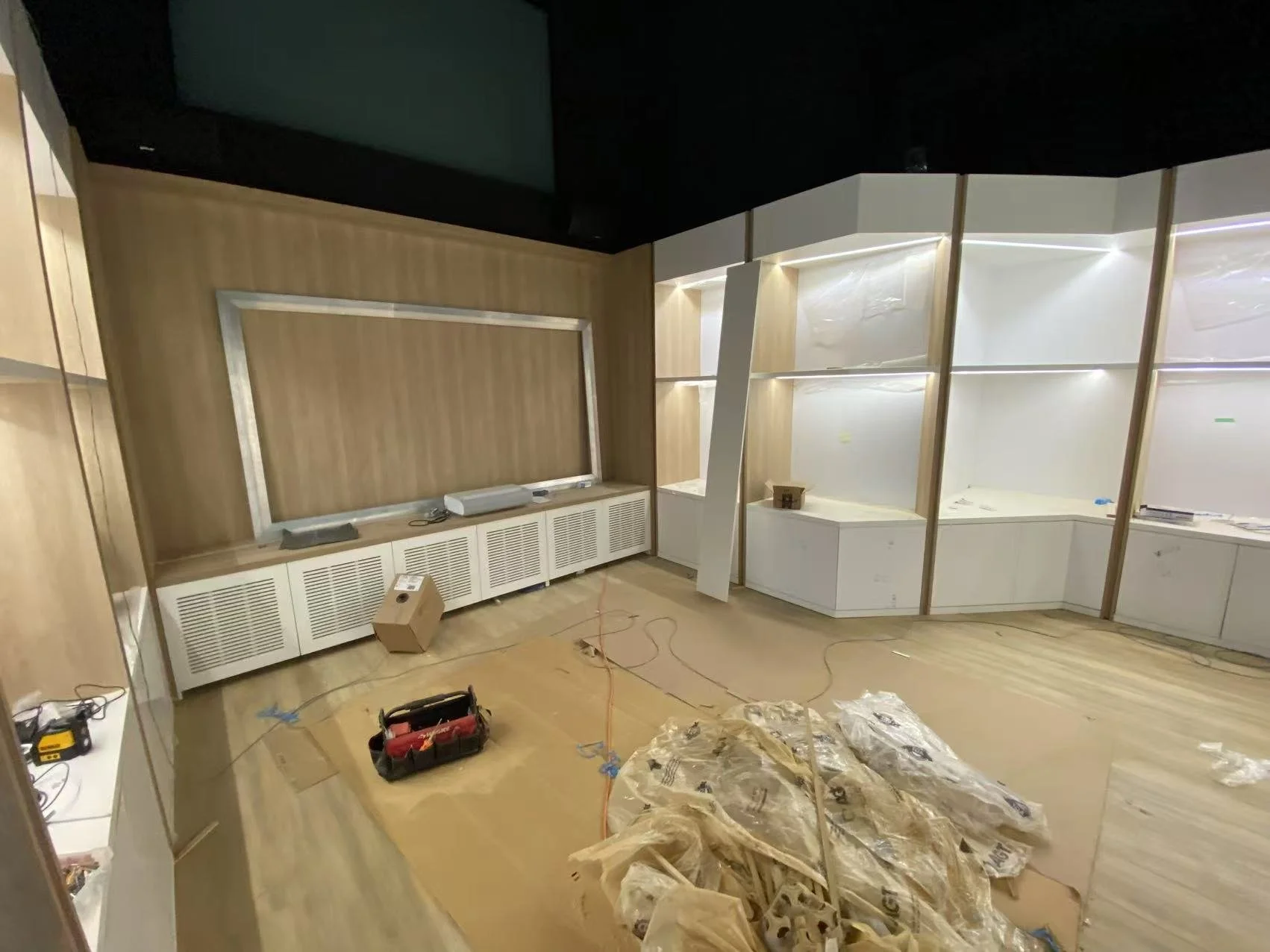 Interior of a room under construction with light wood paneling, white cabinetry, and workspace displays; construction tools and materials are scattered on the floor.