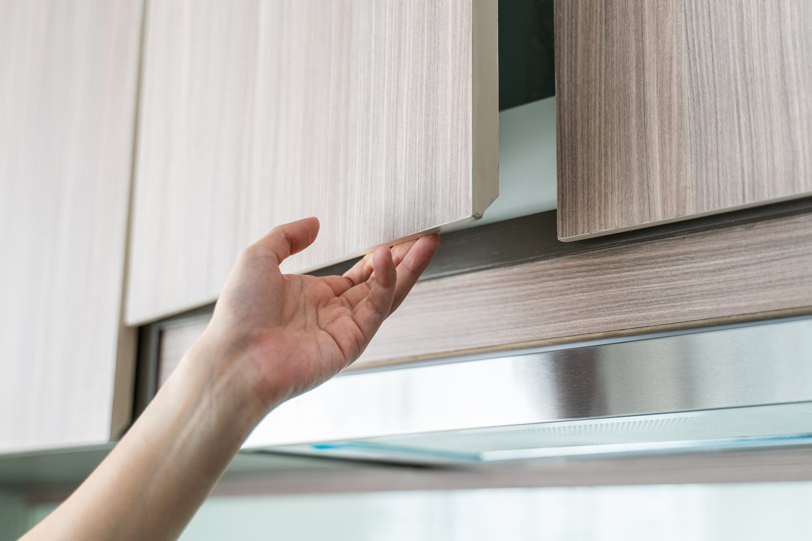 A hand pulling open a light-colored kitchen cabinet door, revealing storage space inside.