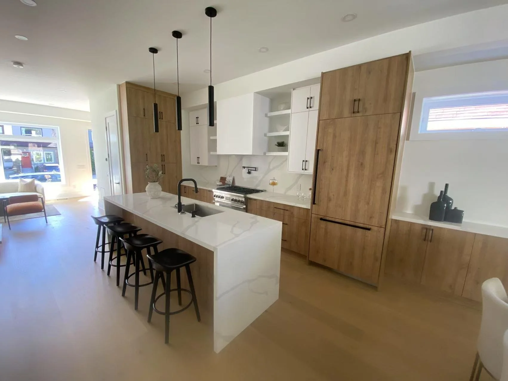 Modern kitchen with white and wood cabinetry, a white marble island with black barstools, black pendant lights, and minimal decor, adjacent to a living area with large windows.