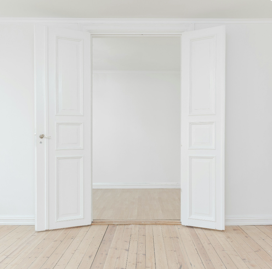 Empty white room with double white doors open to another room with light wood flooring.