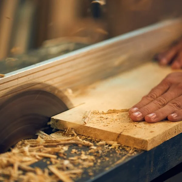 Hands using a circular saw to cut a piece of wood, with wood shavings underneath.