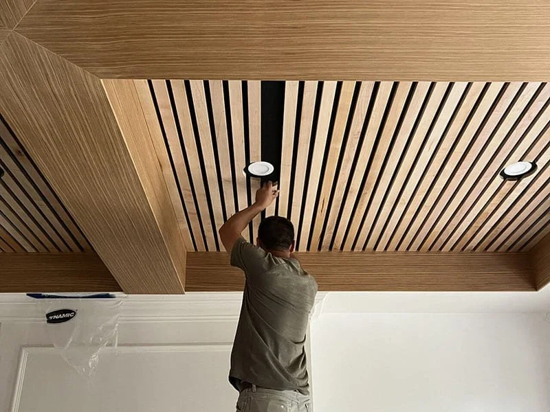 A man installing or adjusting a round recessed ceiling light in a wooden ceiling with vertical black and light wood slats.