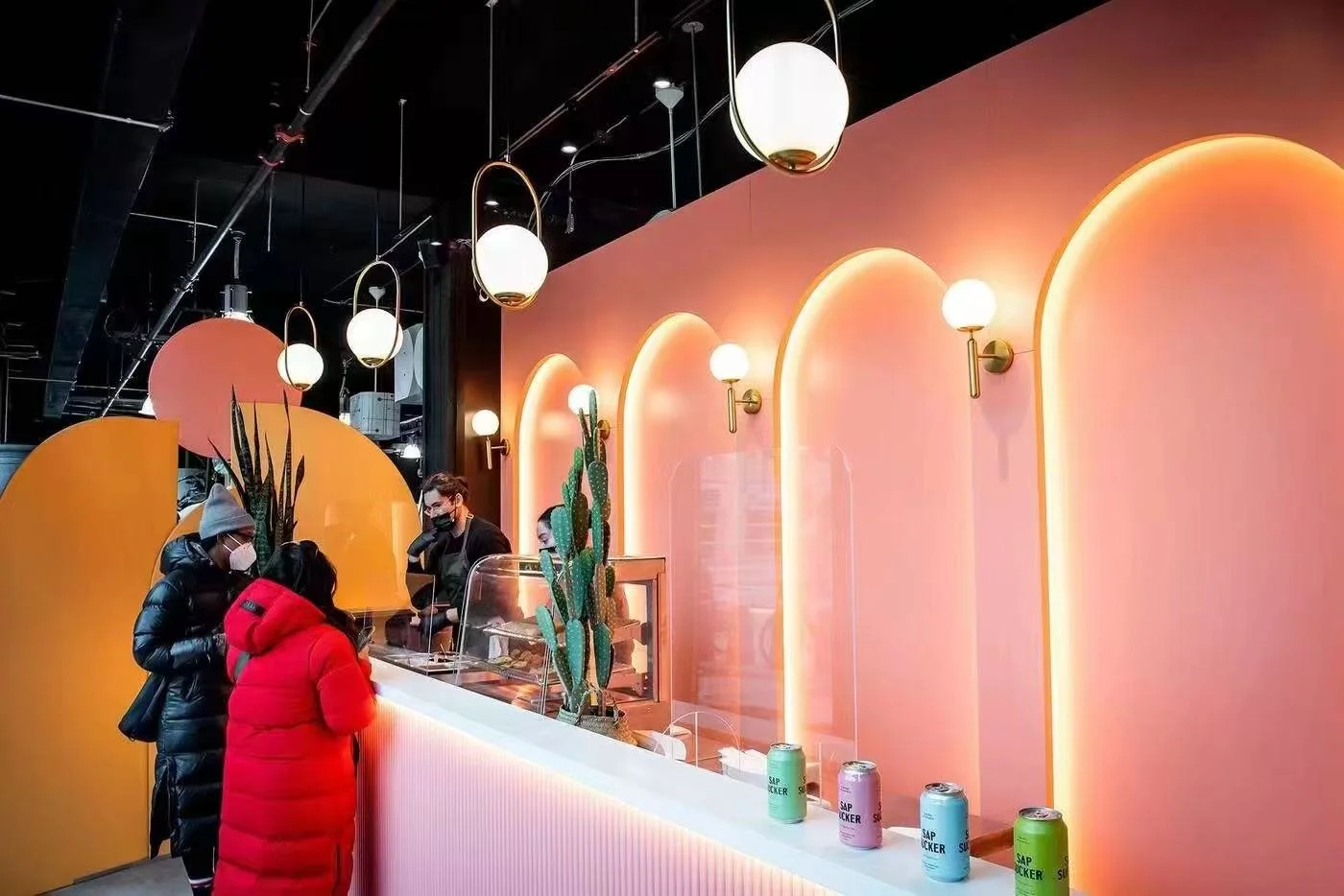 People ordering at a colorful modern cafe counter with pink and orange wall, cactus plants, and hanging spherical lights.