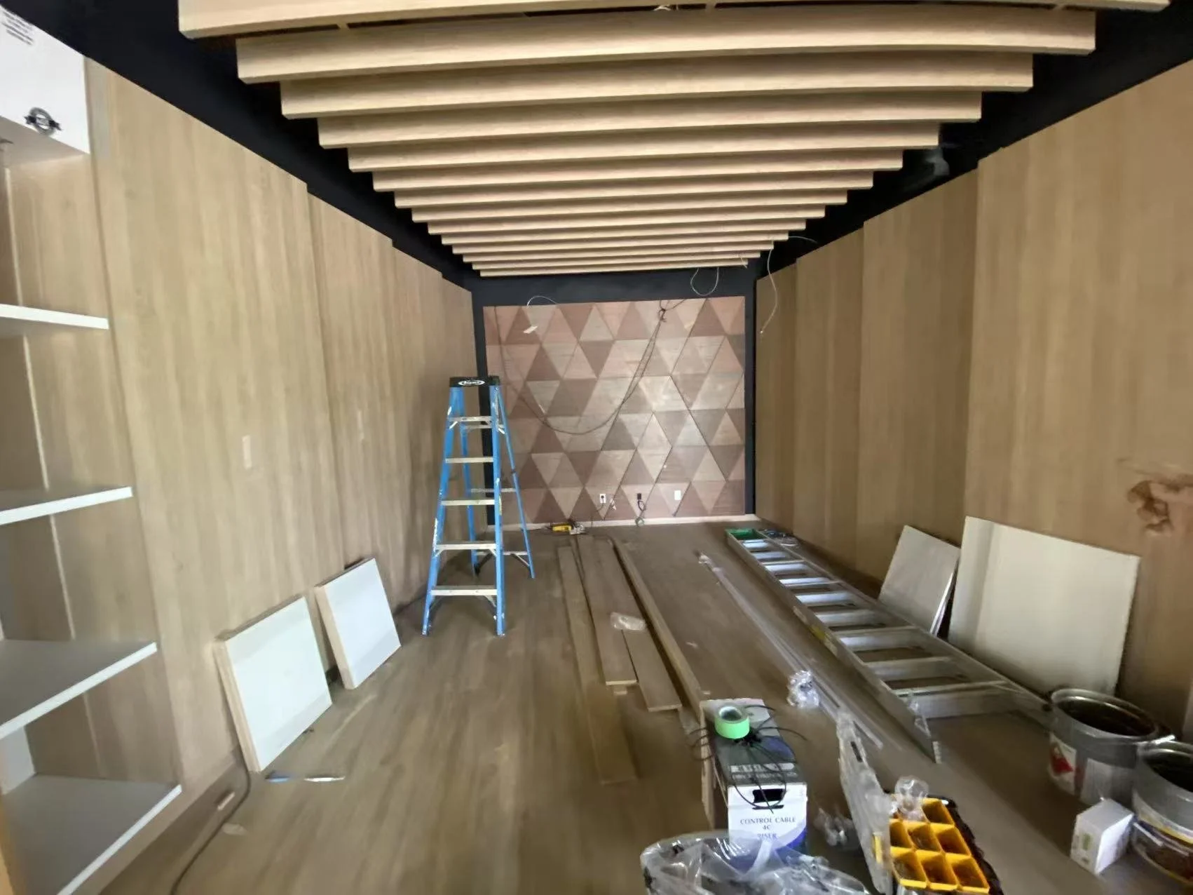 Interior of a room under construction with wooden wall paneling, a blue stepladder, and construction tools and materials on the floor, including a ladder and wires hanging from the ceiling.