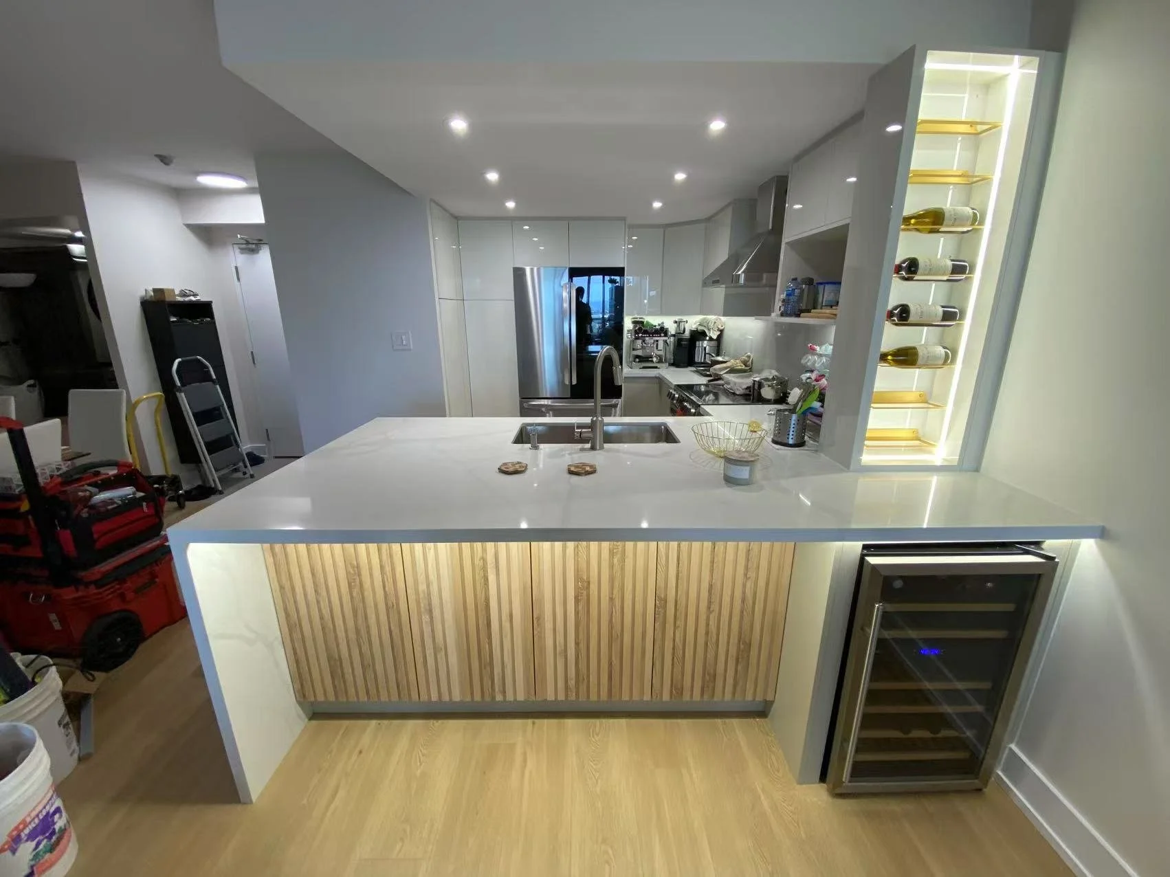 Modern kitchen with a white countertop island, wine rack, stainless steel refrigerator, and built-in wine cooler, with kitchen appliances and supplies on the counters.