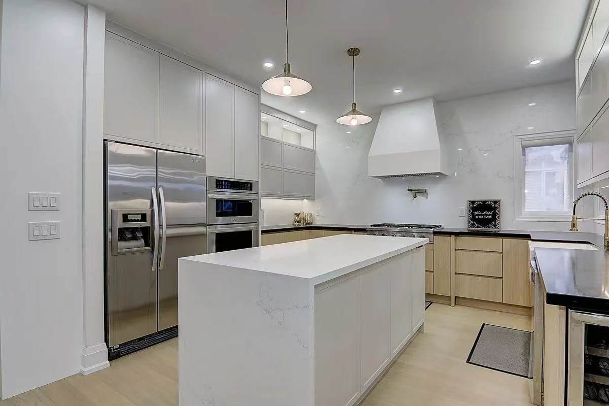 Modern kitchen with white cabinets, stainless steel refrigerator, double oven, white island, black countertop, and white marble backsplash, with a window and ceiling lights.