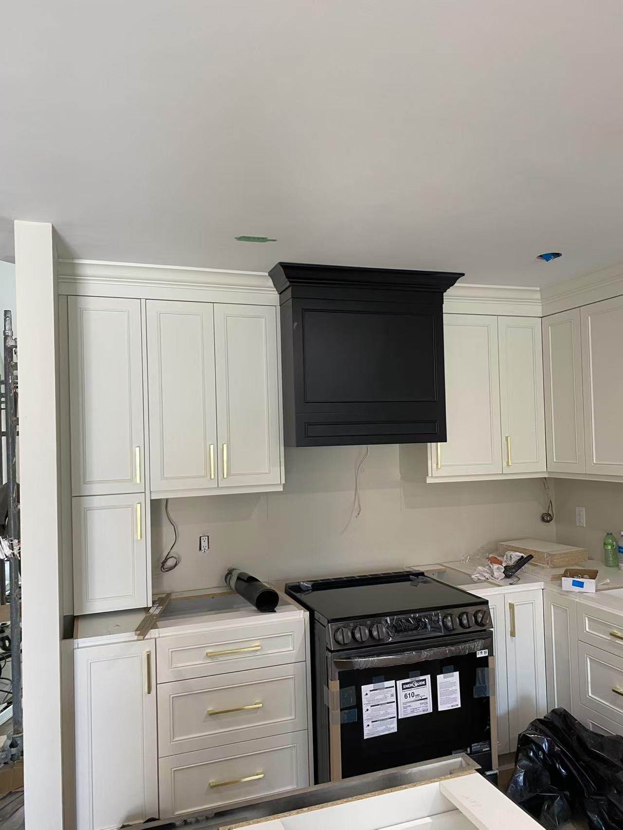 Kitchen under construction with white cabinets, black range hood, black stove, and some tools and materials on the counter.