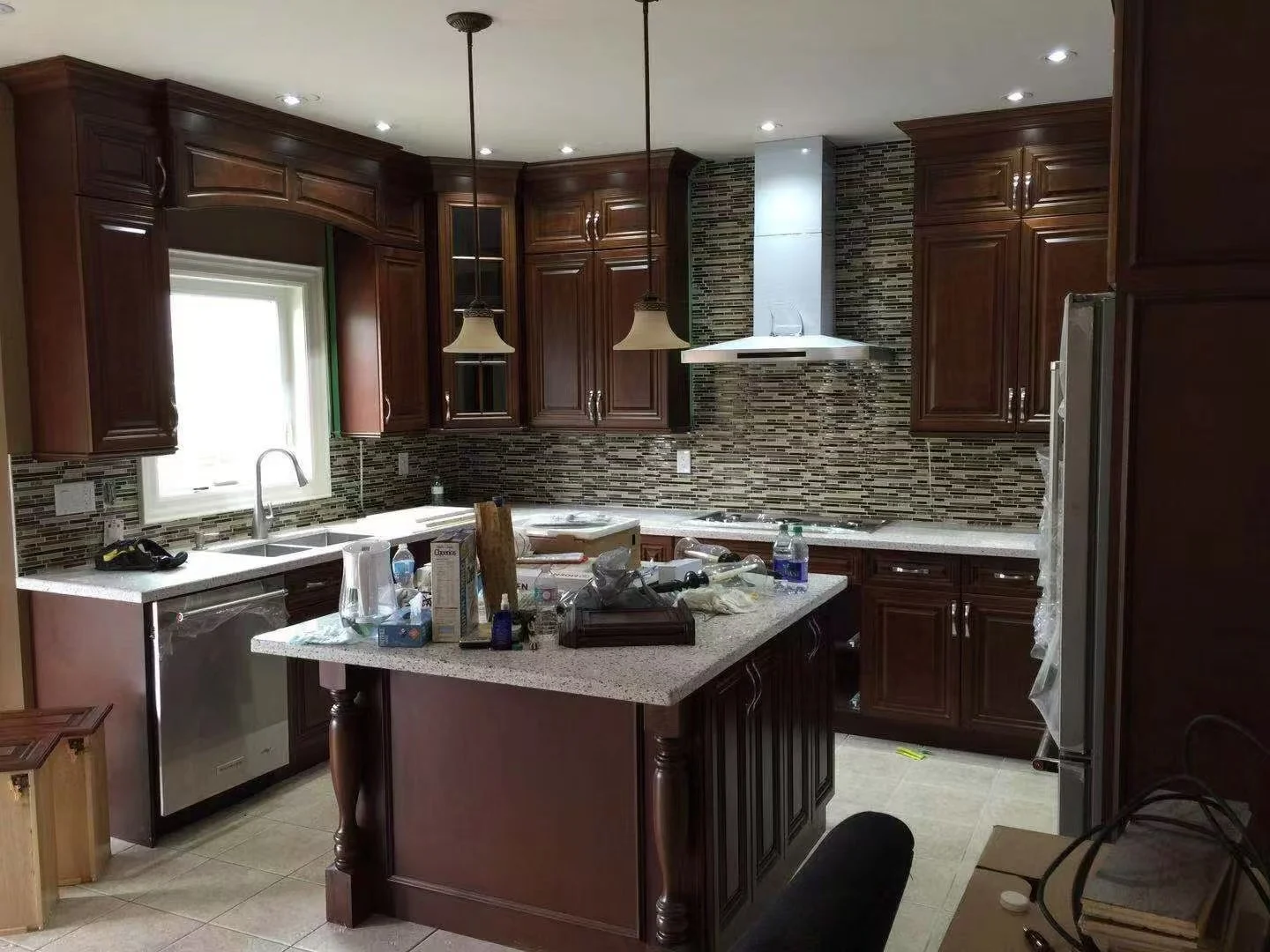 A kitchen with dark wood cabinets, granite countertops, and a mosaic tile backsplash. There is a window above the sink, a stove with a range hood, a dishwasher, and a kitchen island cluttered with various items. Two pendant lights hang over the islan