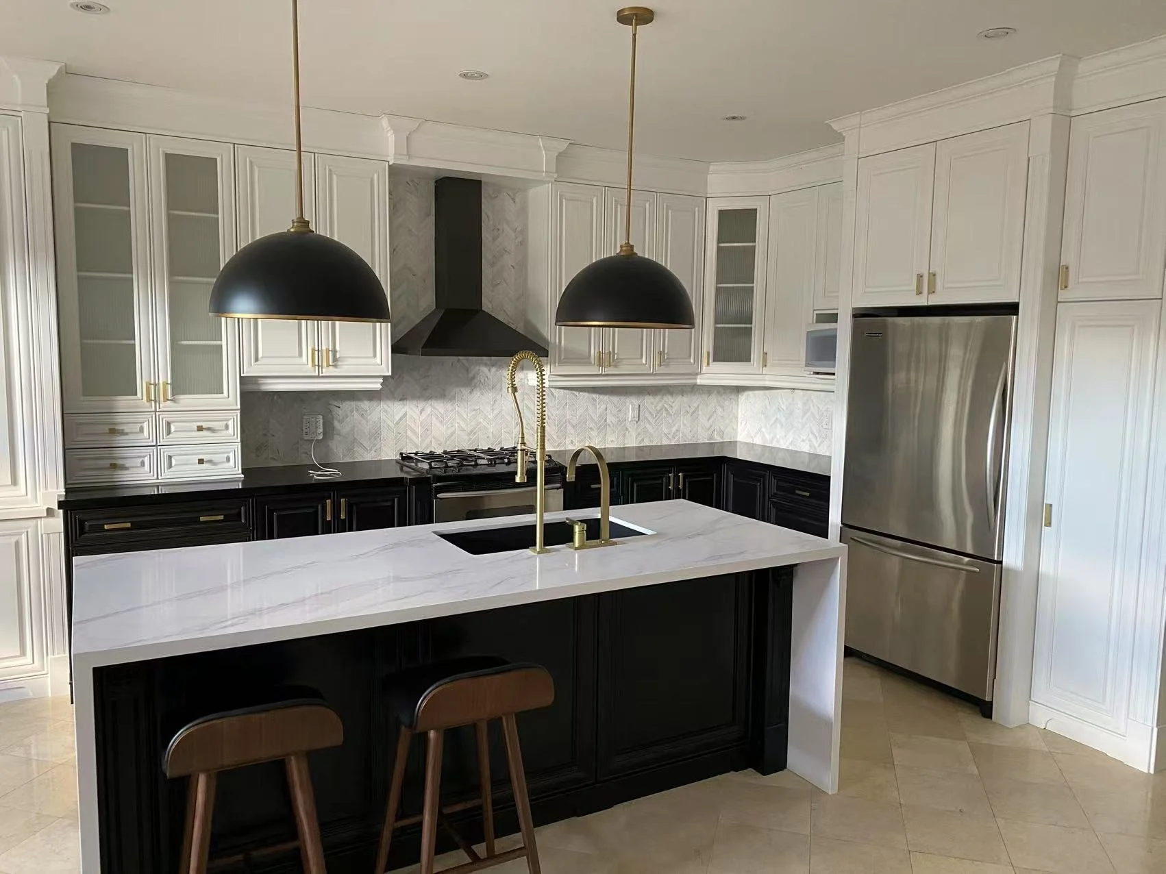Modern kitchen with black and white cabinetry, a central white island with black lower cabinets, gold fixtures, and pendant lights, stainless steel refrigerator, and marble backsplash.