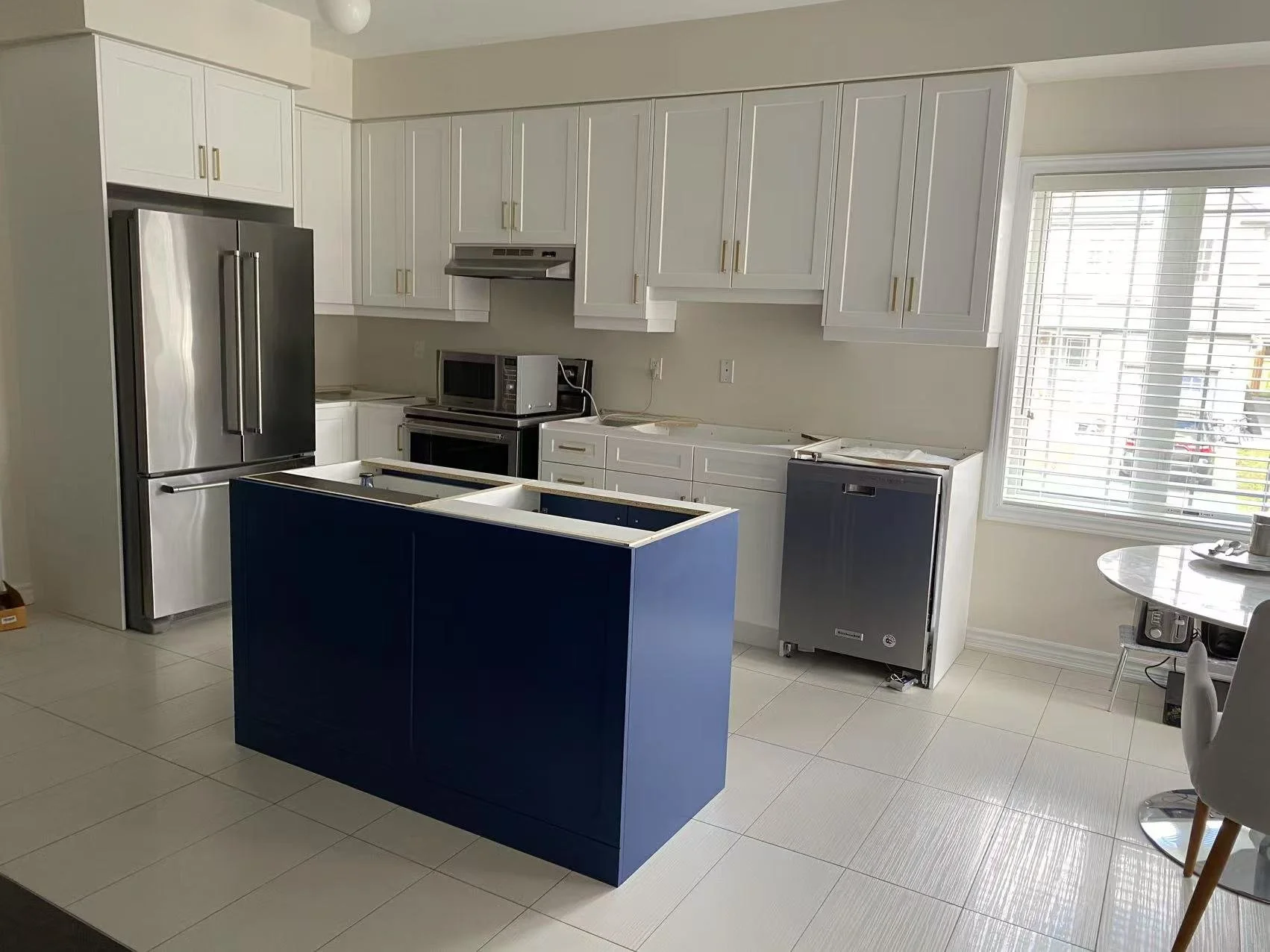 Kitchen with white cabinets, stainless steel refrigerator, microwave, black oven, empty kitchen island with blue front, dishwasher, and window with white blinds.