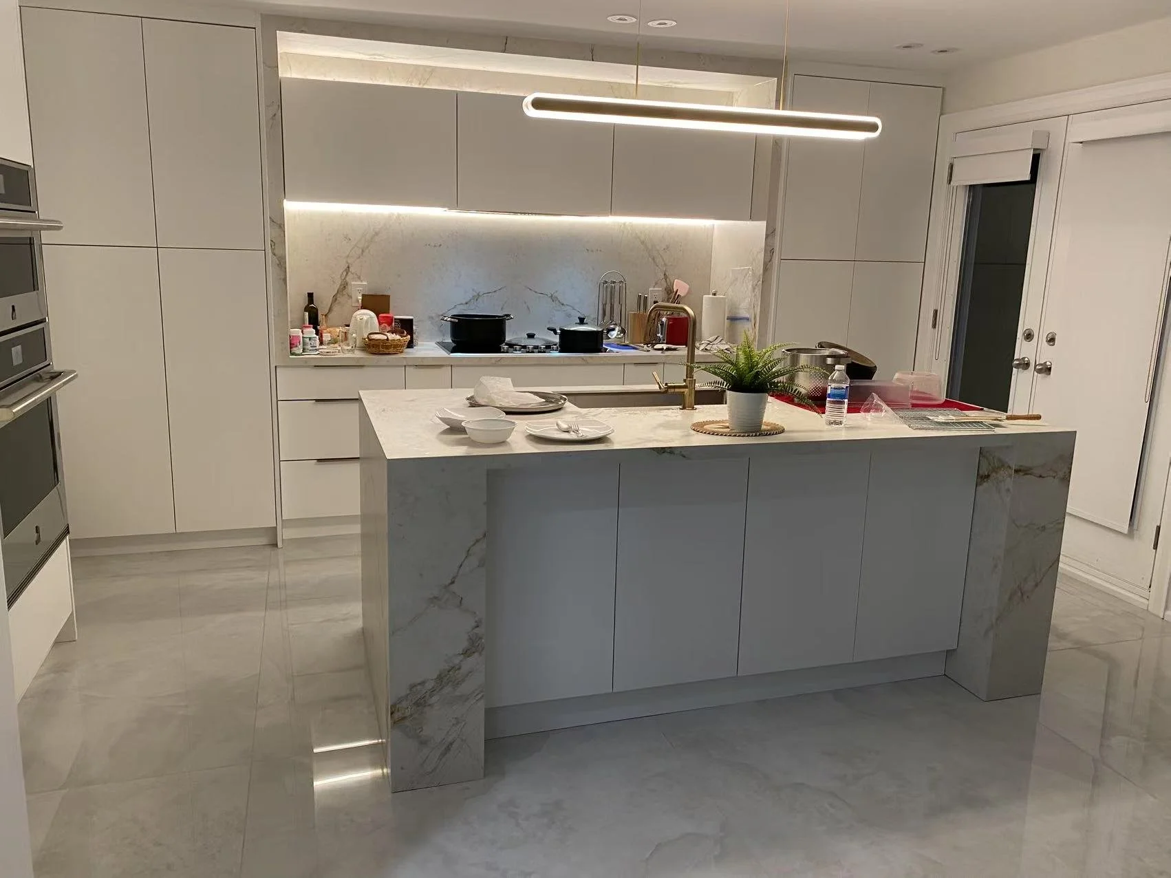 Modern kitchen with white cabinets, marble countertops, and an island. The island has place settings, a potted plant, and a water bottle. The backsplash is marble, with a row of upper cabinets above. Appliances and cooking utensils are visible, with 