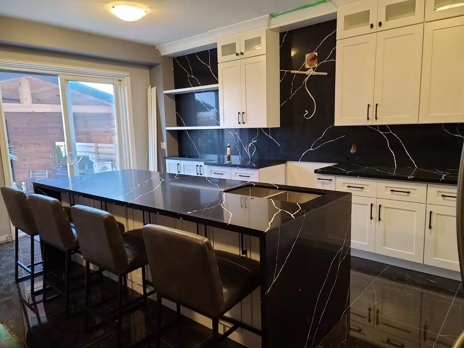 Modern kitchen with white cabinets, black marble countertops and backsplash, black marble island with bar stools, sliding glass door leading to outside.