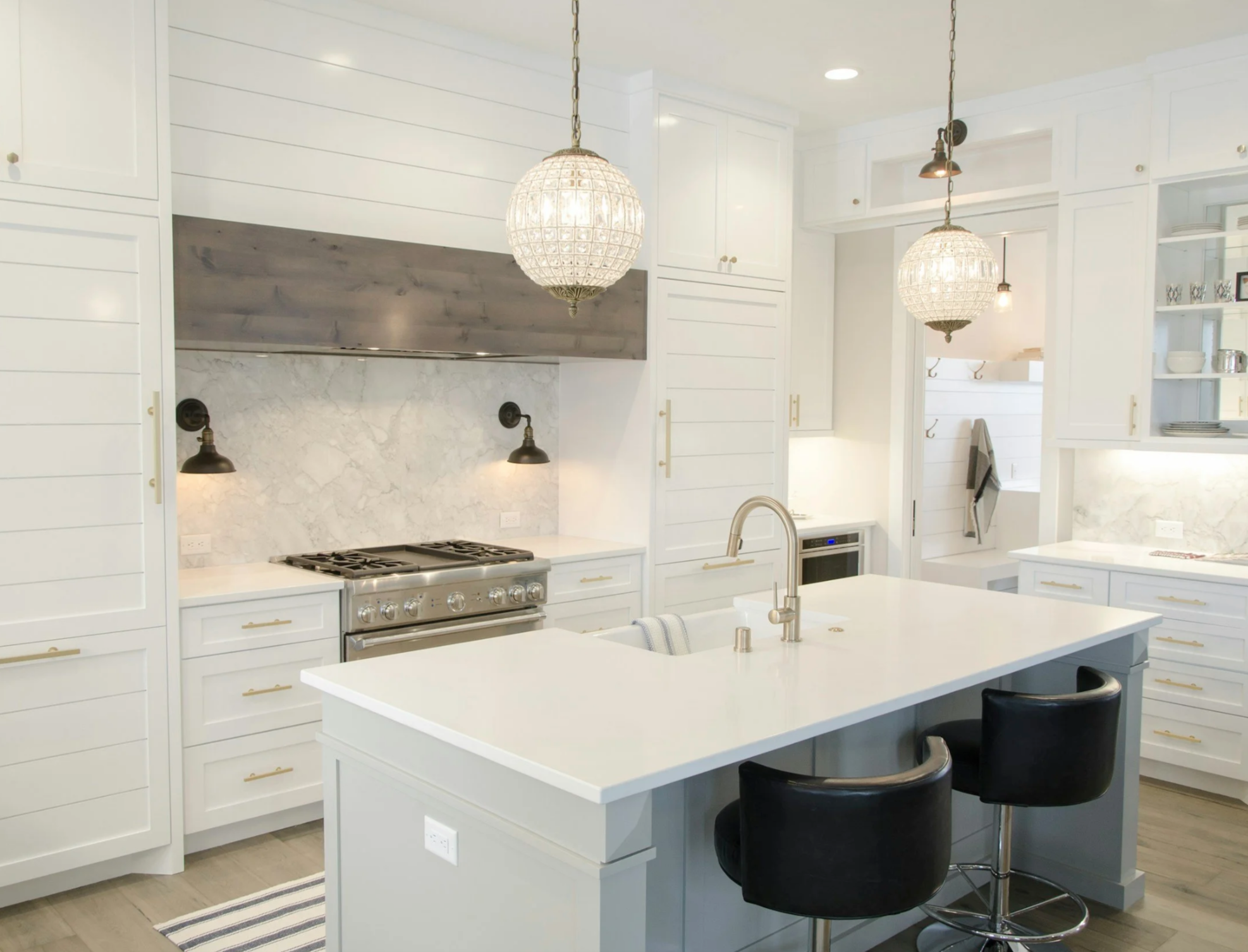 A modern white kitchen with a central island, black chairs, hanging light fixtures, and stainless steel appliances, including a stove and oven, with a marble backsplash and open shelving.