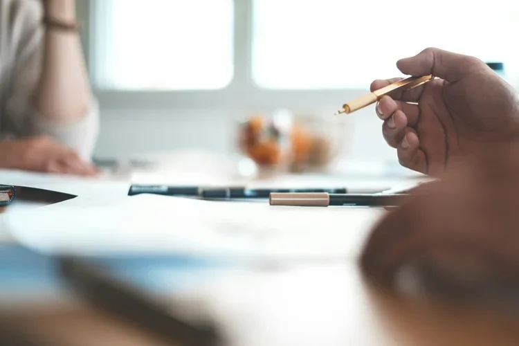 A person holding a pen over a blank sheet of paper on a desk, with other pens and blurred objects in the background.