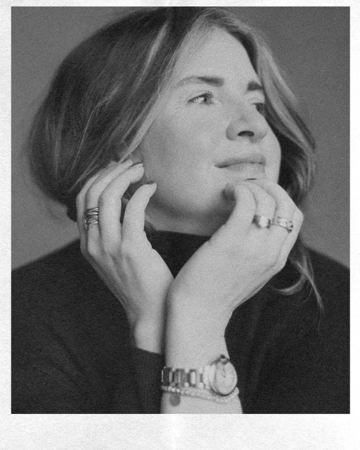 A black and white portrait of a woman with shoulder-length hair, wearing a watch and rings, resting her hand on her chin and neck, looking slightly upward and to the side.