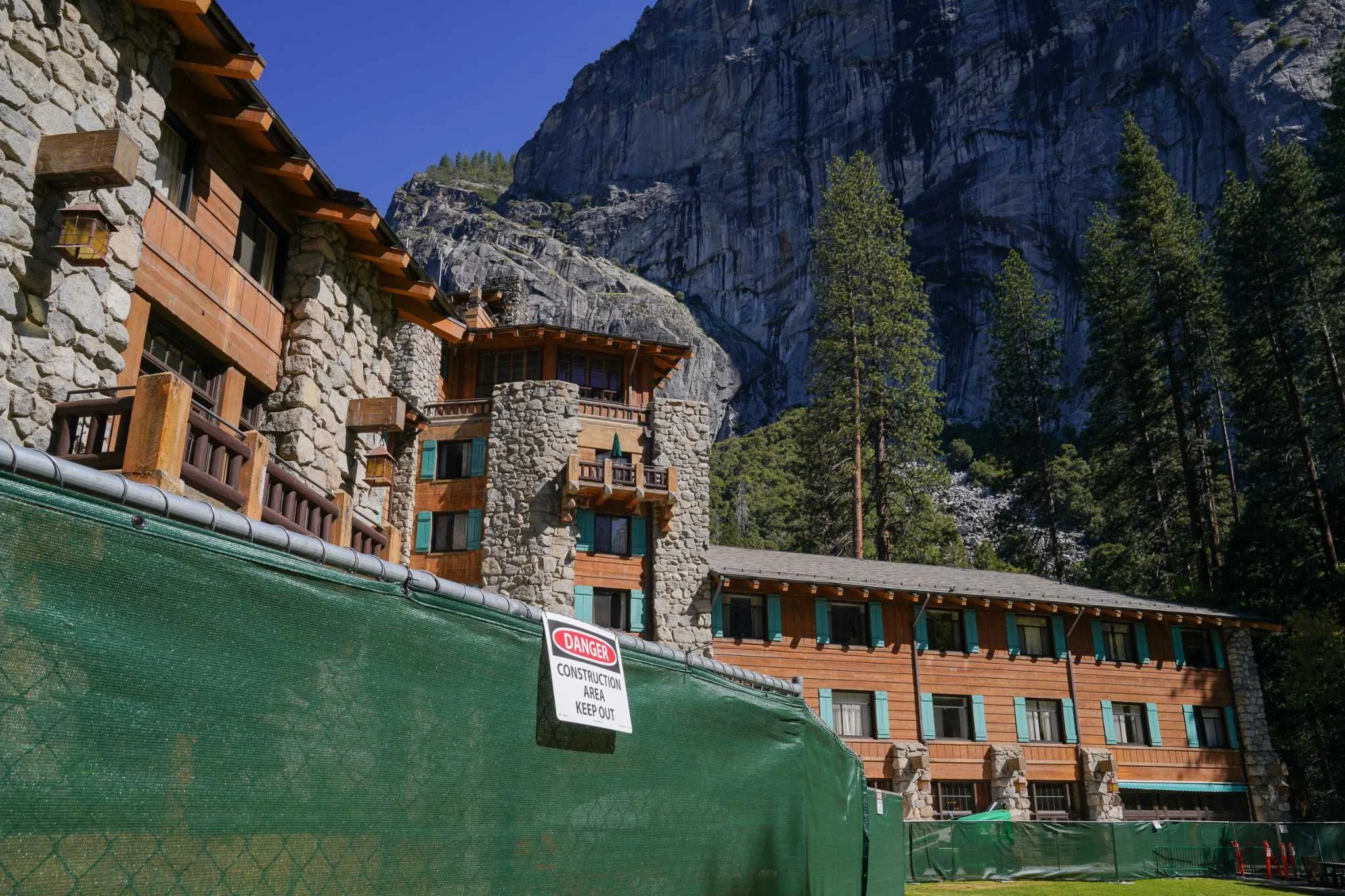 A large stone and wood lodge at a construction site with a green fence and a danger sign, set against a backdrop of tall pine trees and rocky mountain cliffs under a clear blue sky.