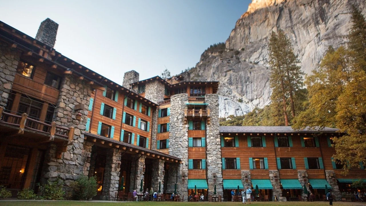 A large mountain lodge made of stone and wood, with a outdoor patio and trees, set against a mountain backdrop.