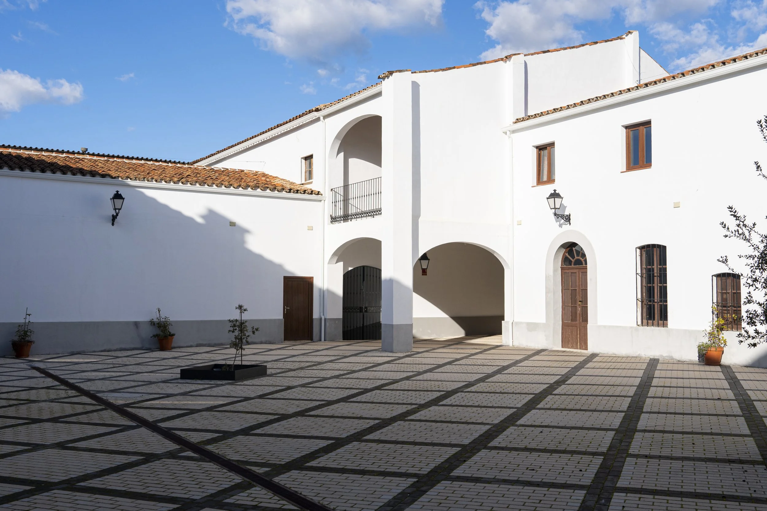 Vista de un patio con piso de ladrillos, paredes blancas, puertas y ventanas de madera, faroles en la pared, y un cielo azul con algunas nubes.