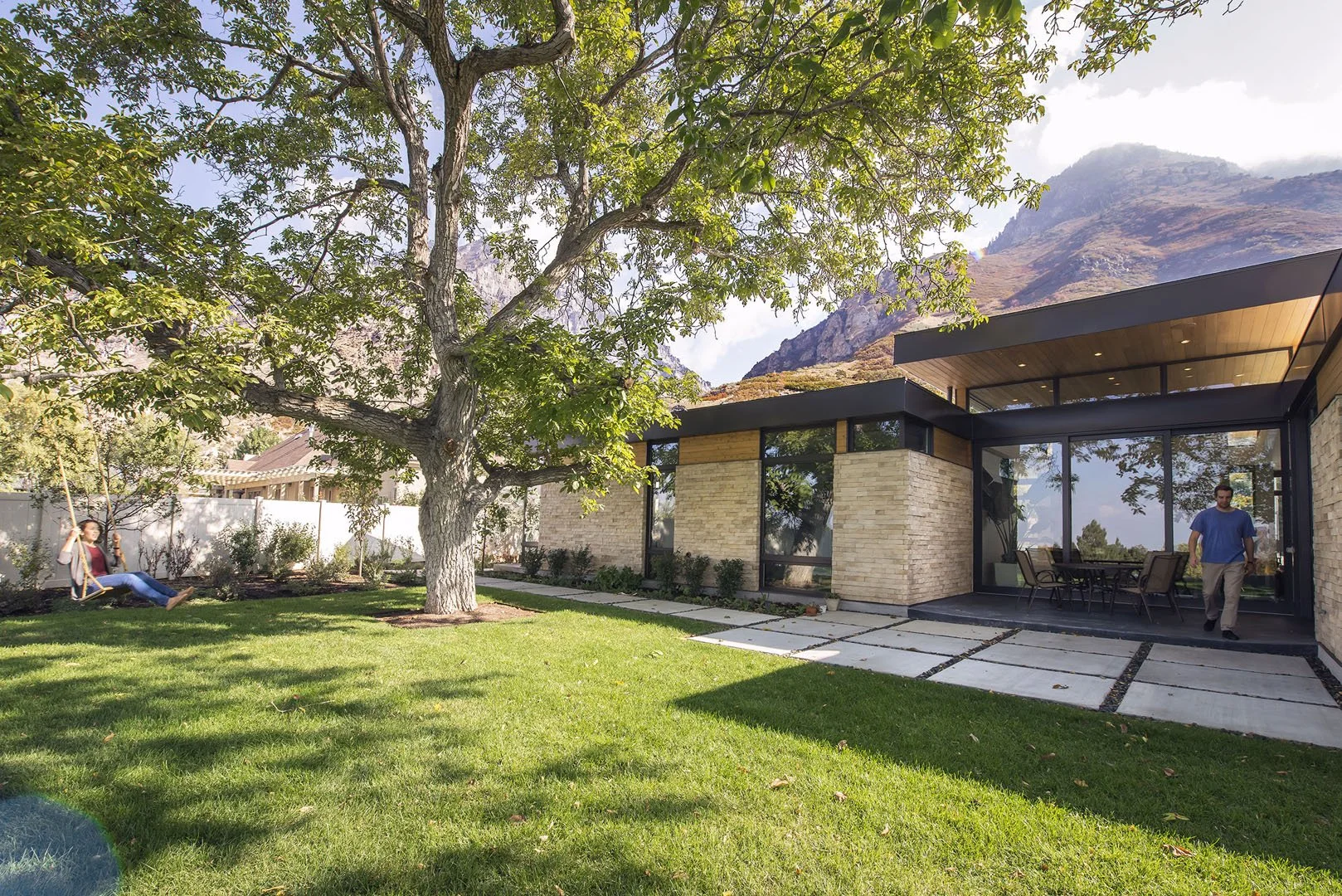 Modern stone-and-wood home with deep overhangs behind a mature shade tree, with mountains beyond, Rock Canyon, Provo, Utah.