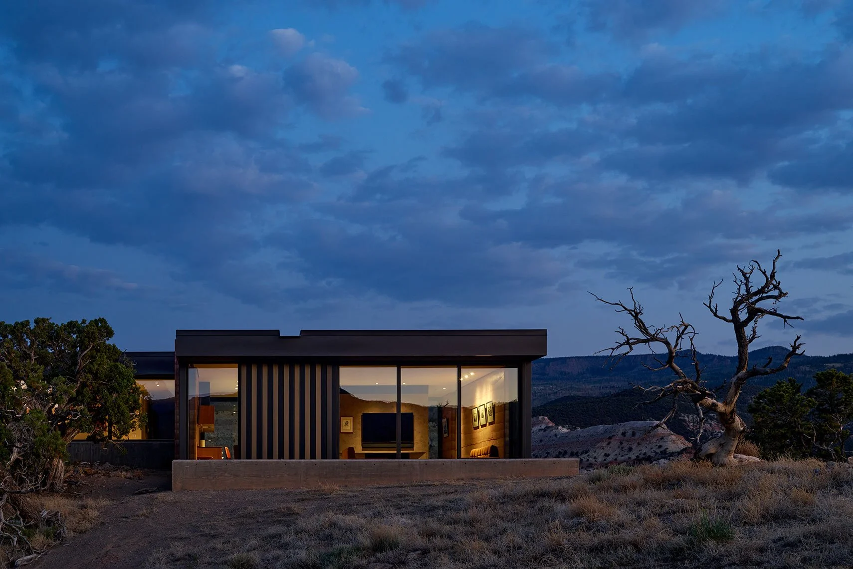Modern desert home glowing at dusk beside a weathered tree, with mountains in the distance, No Neh She, Teasdale, Utah.