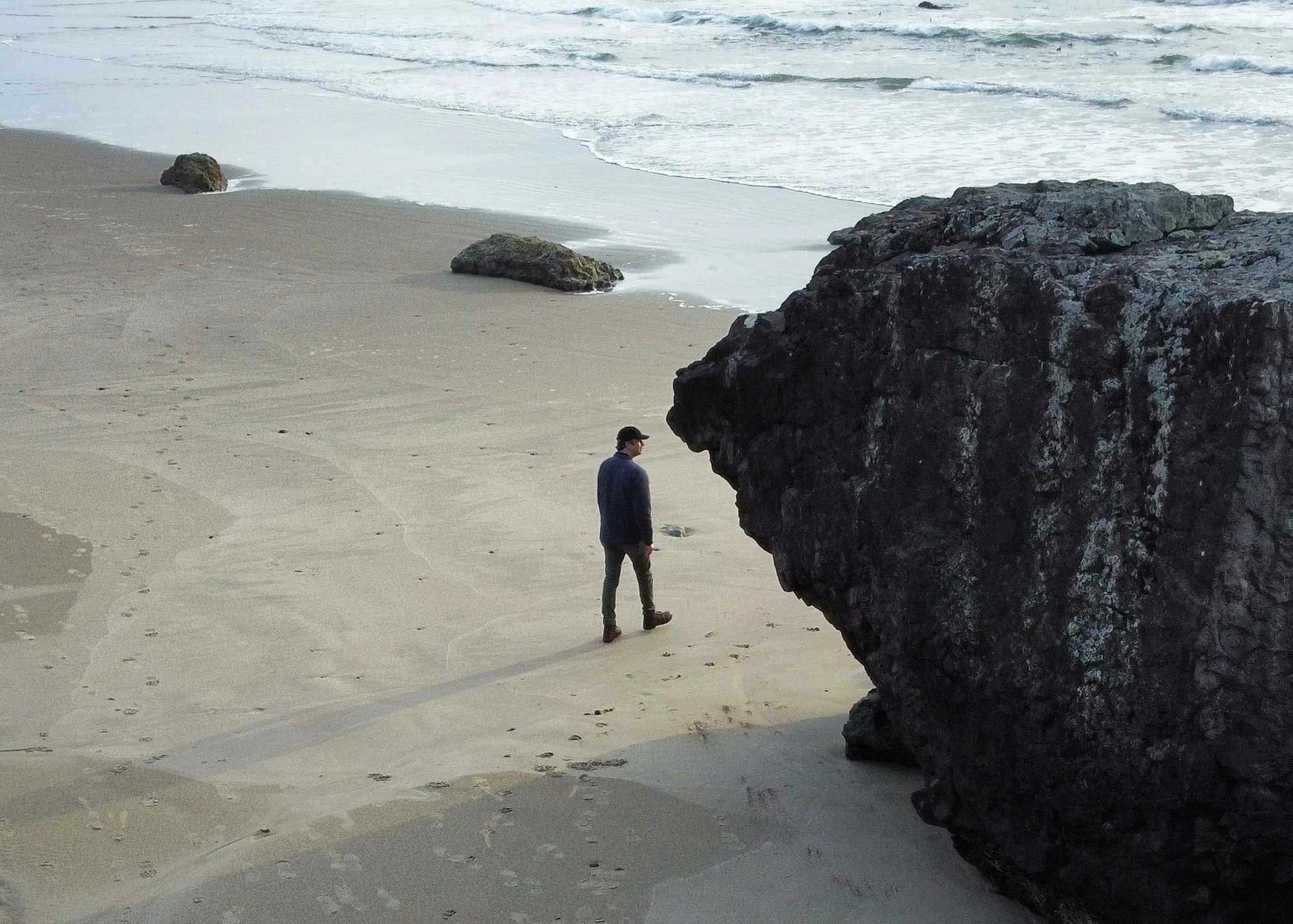 Person walking on a sandy beach near large rocks, with the ocean and waves in the background.