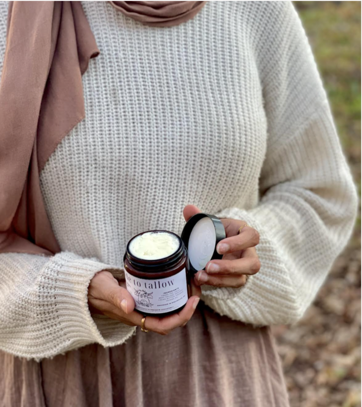 A woman in a cream knitted sweater holds an open jar of white cream or lotion labeled 'to tallow' in an outdoor setting with blurred foliage in the background.