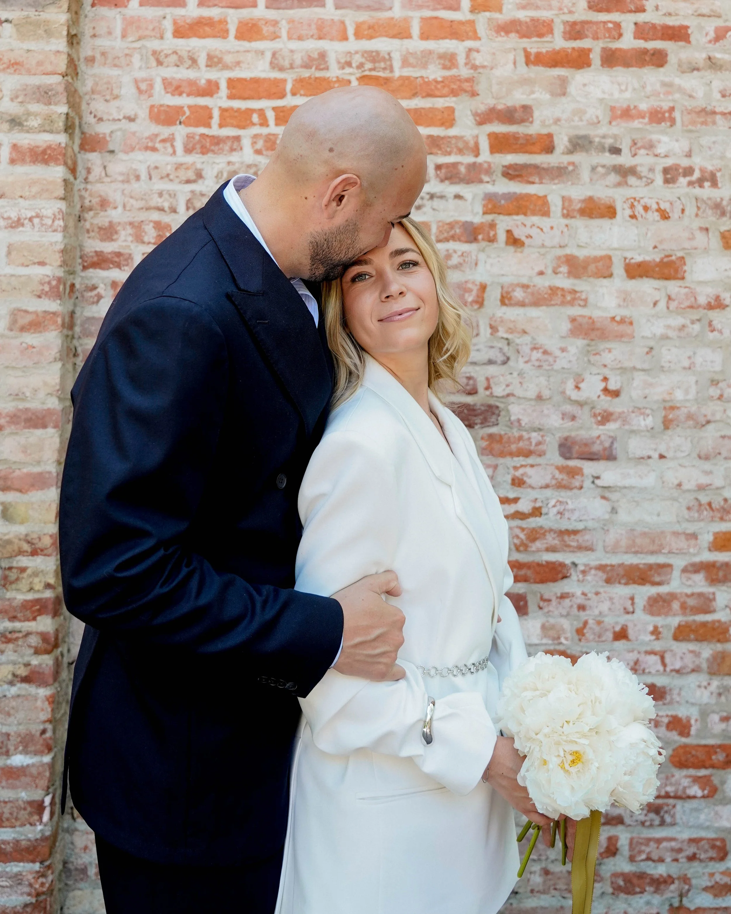 A man in a dark suit gently kisses a woman in a white blazer on the forehead as she holds a bouquet of white flowers, standing against a brick wall.