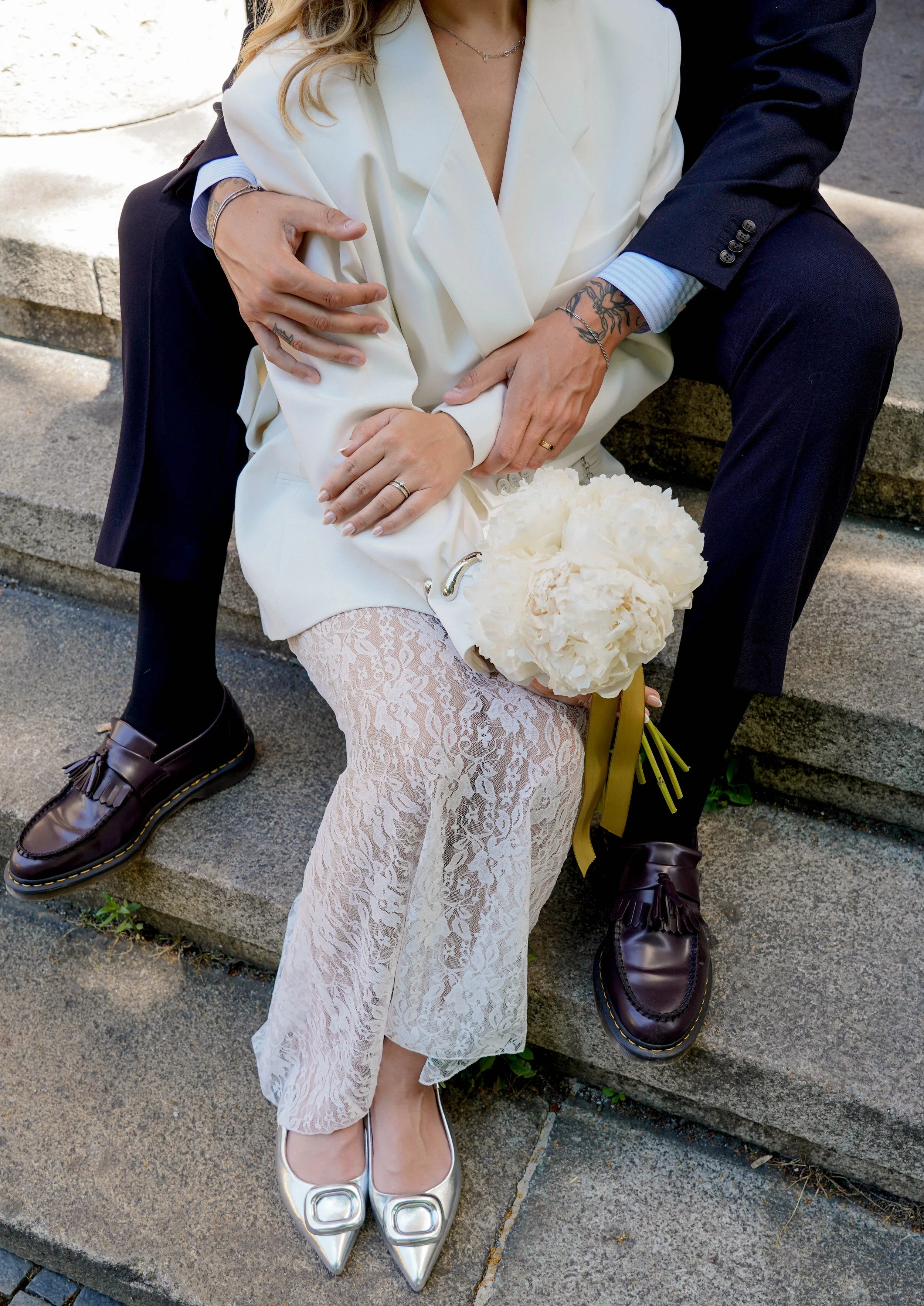 Close-up of a couple sitting on steps, the woman in a white blazer and lace skirt with silver shoes, holding a bouquet of white flowers, and the man in a dark suit with tattoos on his arm, wearing black shoes.