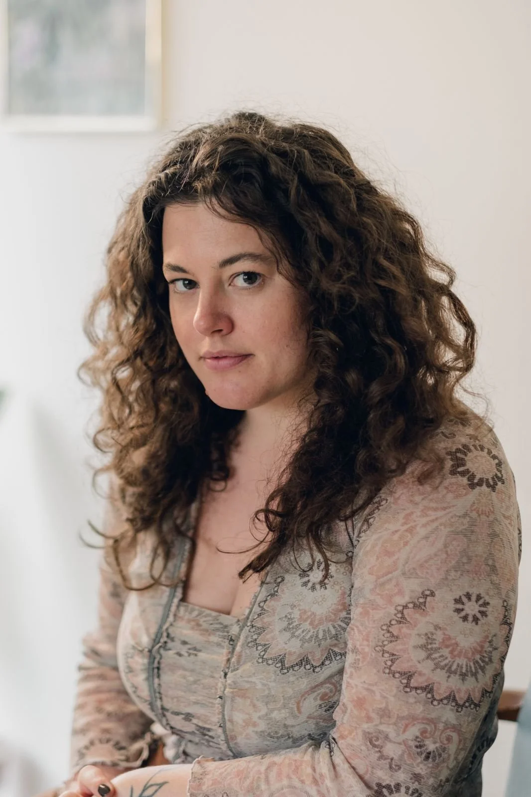 A woman with long, curly brown hair looking at the camera, wearing a patterned long-sleeve top, indoors with a window in the background.