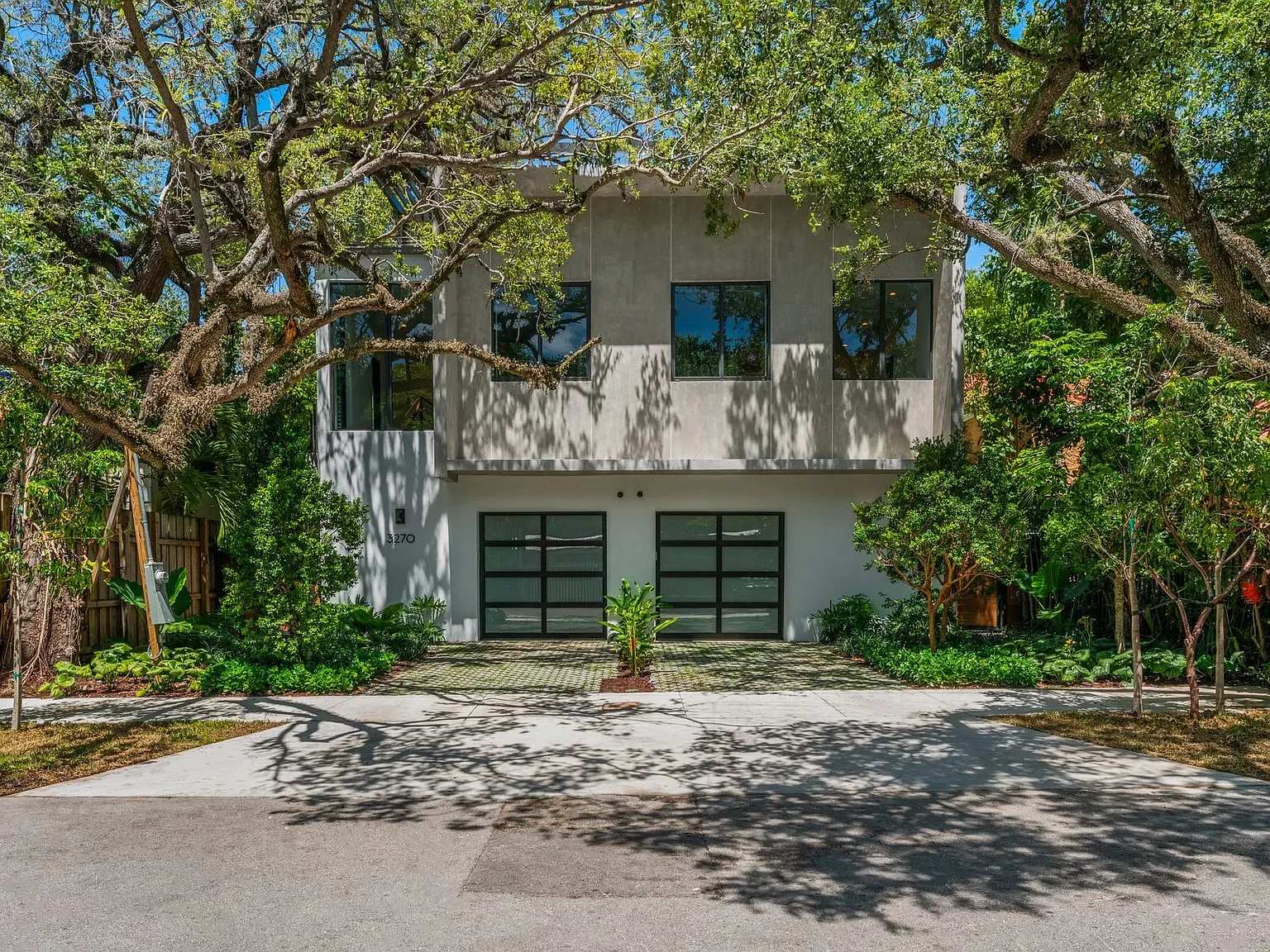 Modern two-story house with a concrete facade, black framed windows, and a glass garage door, surrounded by lush trees and greenery, with shadows cast on the front driveway.