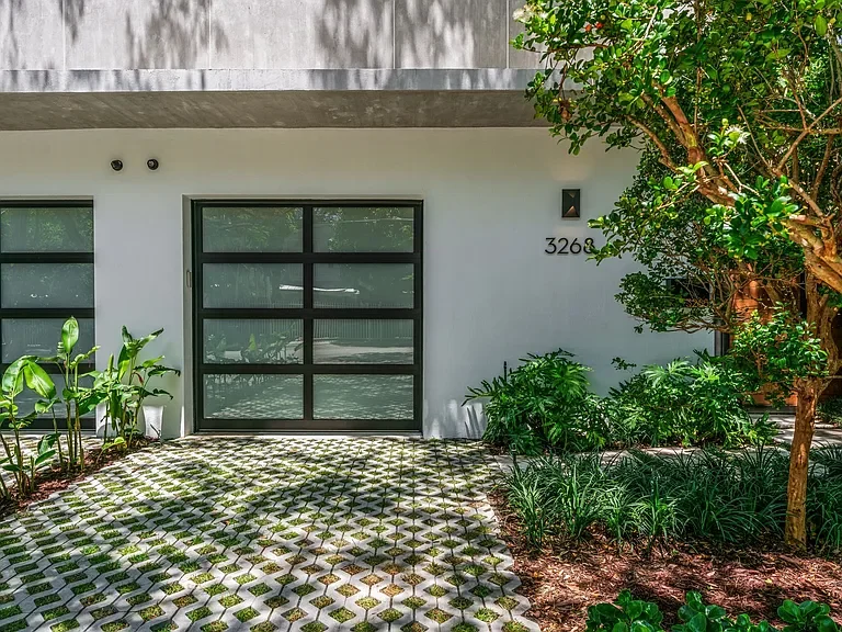 Modern house entrance with black-framed glass door and windows, surrounded by green plants and trees, with a patterned concrete walkway.